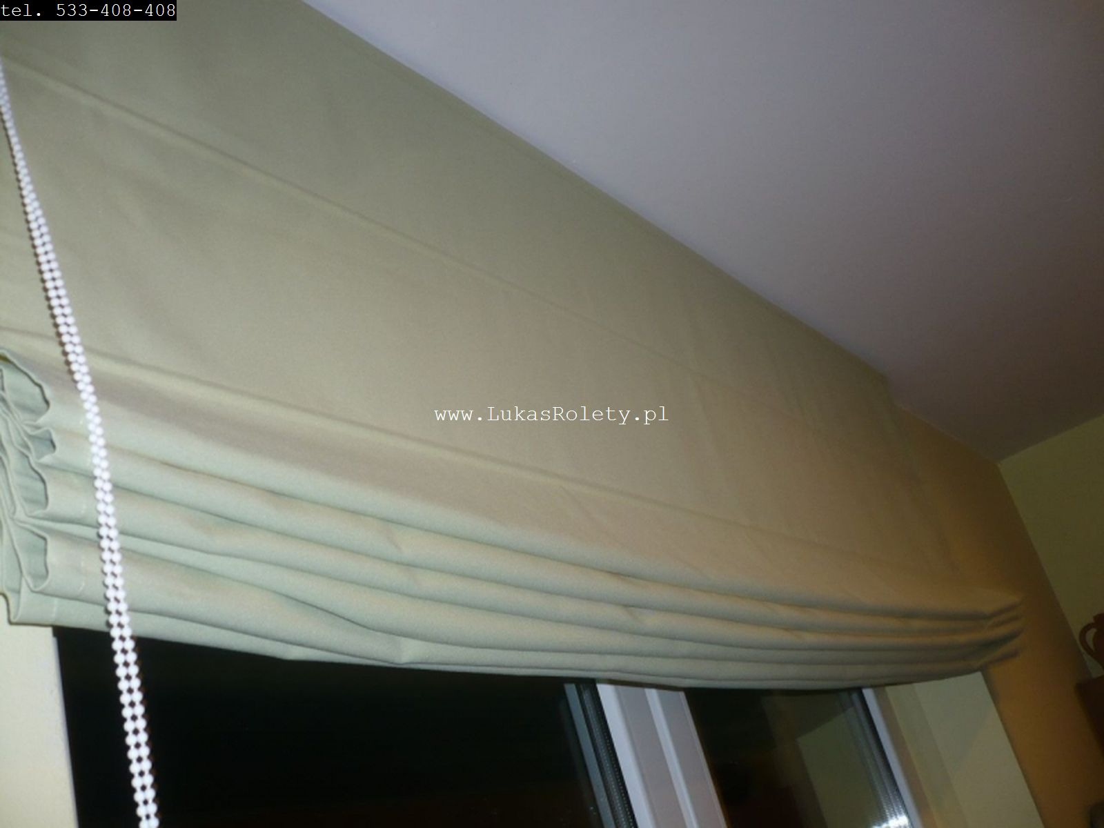 Jasnozielona, częściowo zwinięta tkanina na oknie, sterowana białym łańcuszkiem, widoczna z dołu pod kątem, z adresem strony internetowej na materiale.