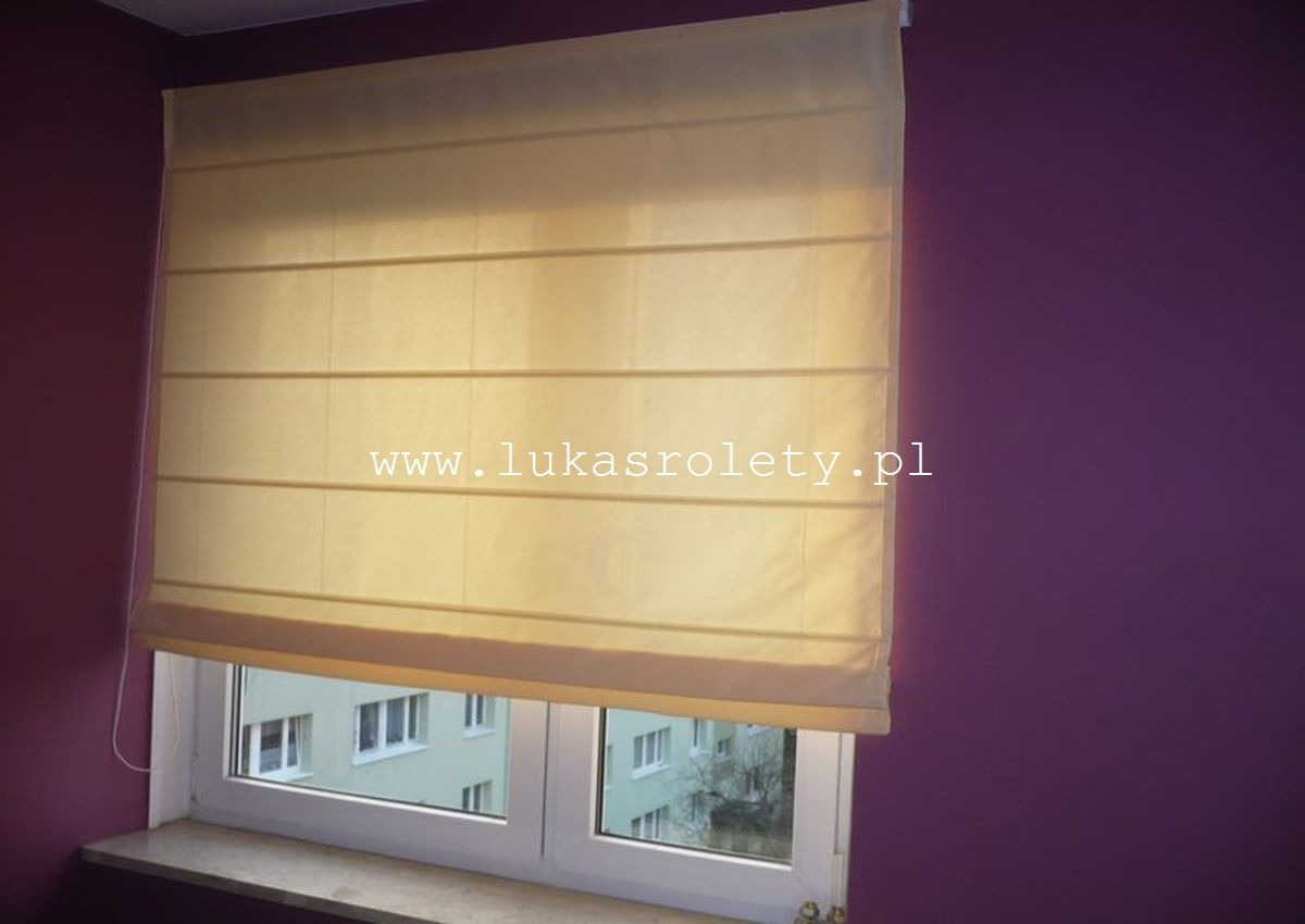 Beżowa tkanina ułożona w poziome fałdy, zamontowana w oknie z widokiem na budynek mieszkalny, fioletowa ściana w tle, widoczny adres strony internetowej na produkcie.