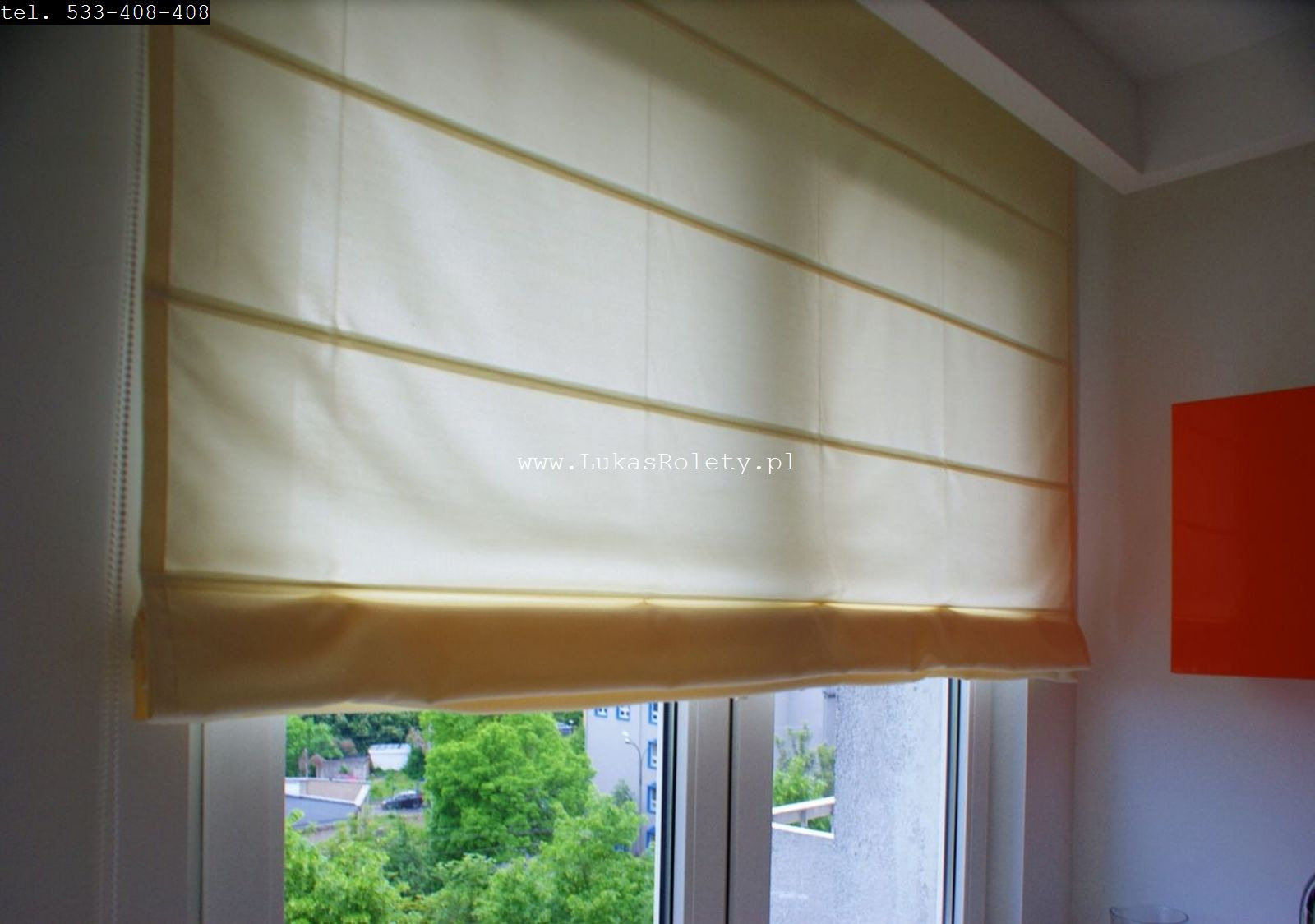 Beżowa, zwinięta tkanina okienna z poziomymi przeszyciami, zamontowana w oknie z widokiem na zieleń miejską.