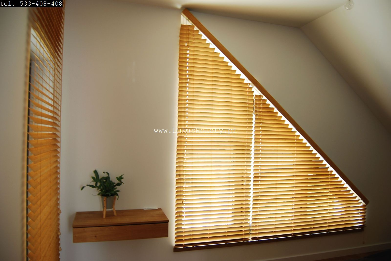 Jasne drewniane żaluzje poziome zamontowane w oknie o nietypowym, trójkątnym kształcie na poddaszu, obok mała drewniana półka z rośliną doniczkową.