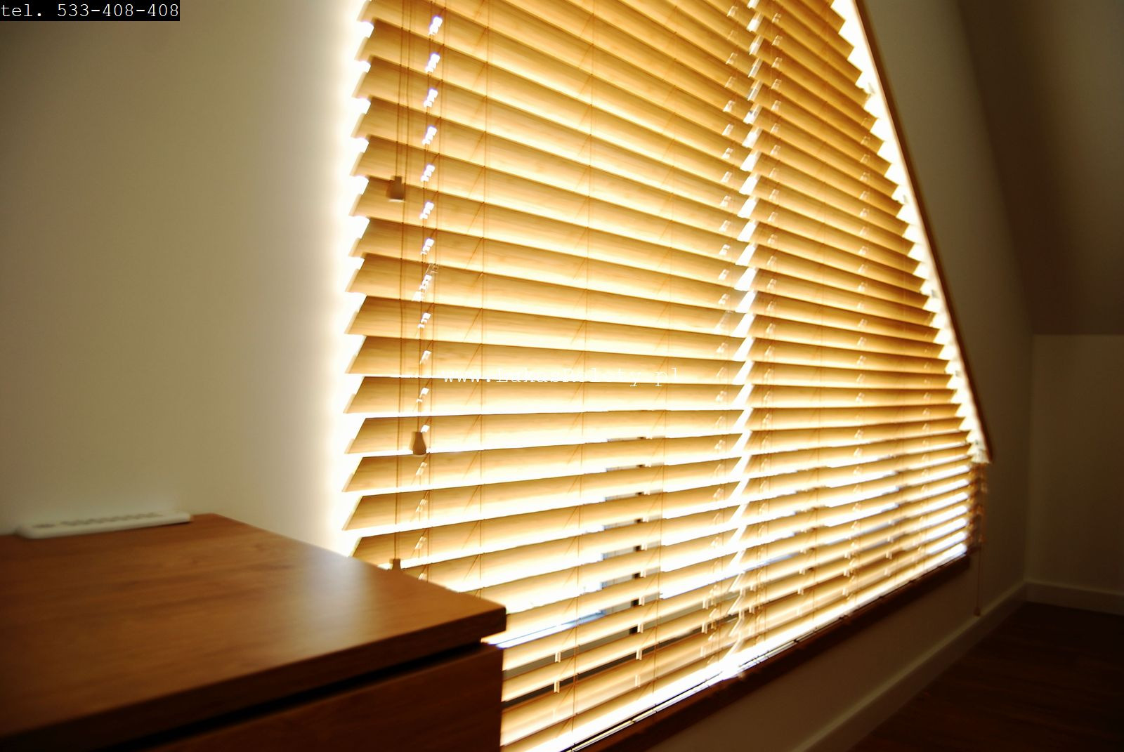 Jasne drewniane lamele żaluzji, z delikatnym oświetleniem od strony okna, częściowo zasłaniają okno w poddaszu, obok widoczny fragment drewnianej komody.