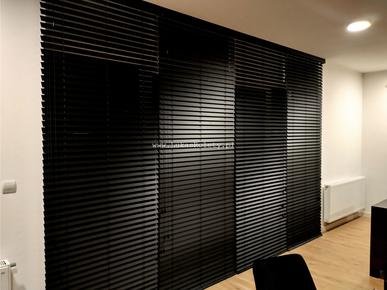 Ciemne, poziome lamele żaluzji drewnianych zamontowanych w oknach, widoczne z wnętrza pomieszczenia. Na lamelach widoczny adres strony internetowej.