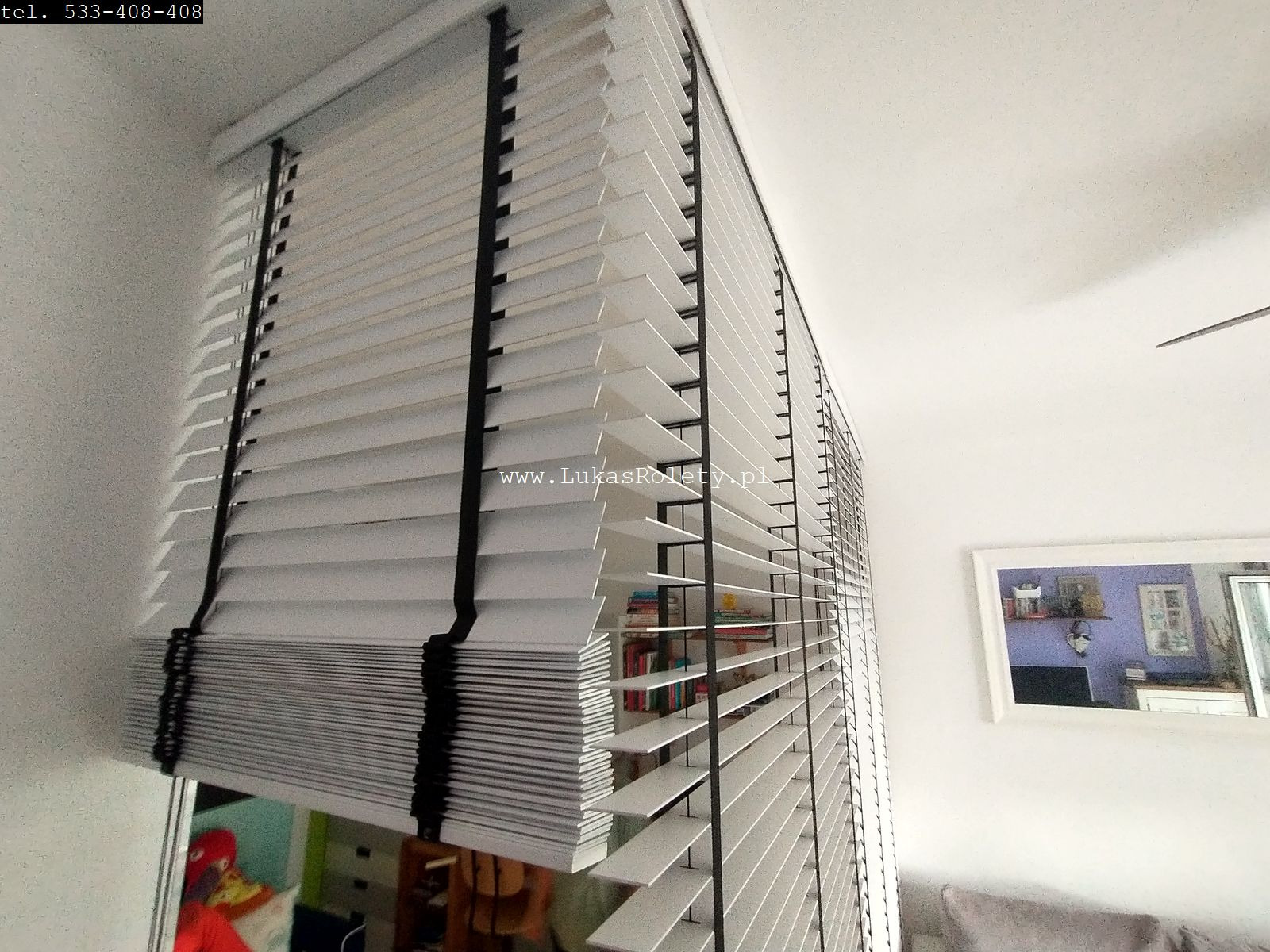 Białe żaluzje drewniane z czarnymi taśmami, częściowo podciągnięte, widok z dołu ukazujący strukturę lameli i elementy montażowe, z widocznym odbiciem wnętrza pokoju w szybie okna.