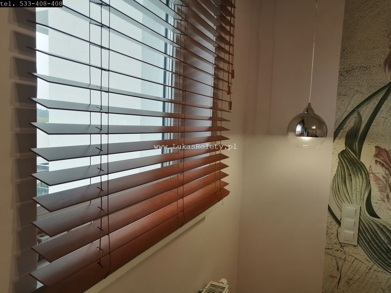 Ciemnobrązowe żaluzje drewniane w oknie, częściowo zasłonięte, z widocznym mechanizmem sznurkowym, w tle ściana z kwiatowym motywem i nowoczesna lampa wisząca.