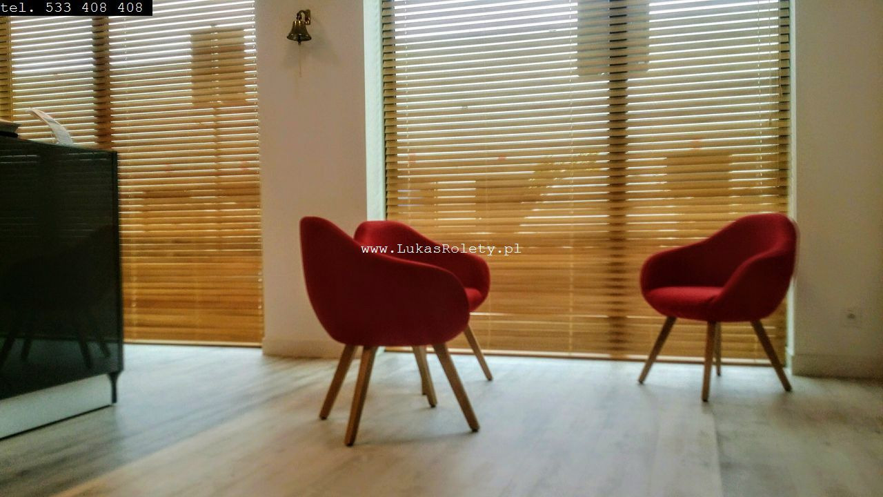 Wnętrze pokoju z drewnianymi żaluzjami poziomymi w kolorze jasnego drewna, dwa czerwone krzesła na drewnianych nogach, widoczny fragment czarnego mebla z lewej strony, na ścianie złoty dzwonek.