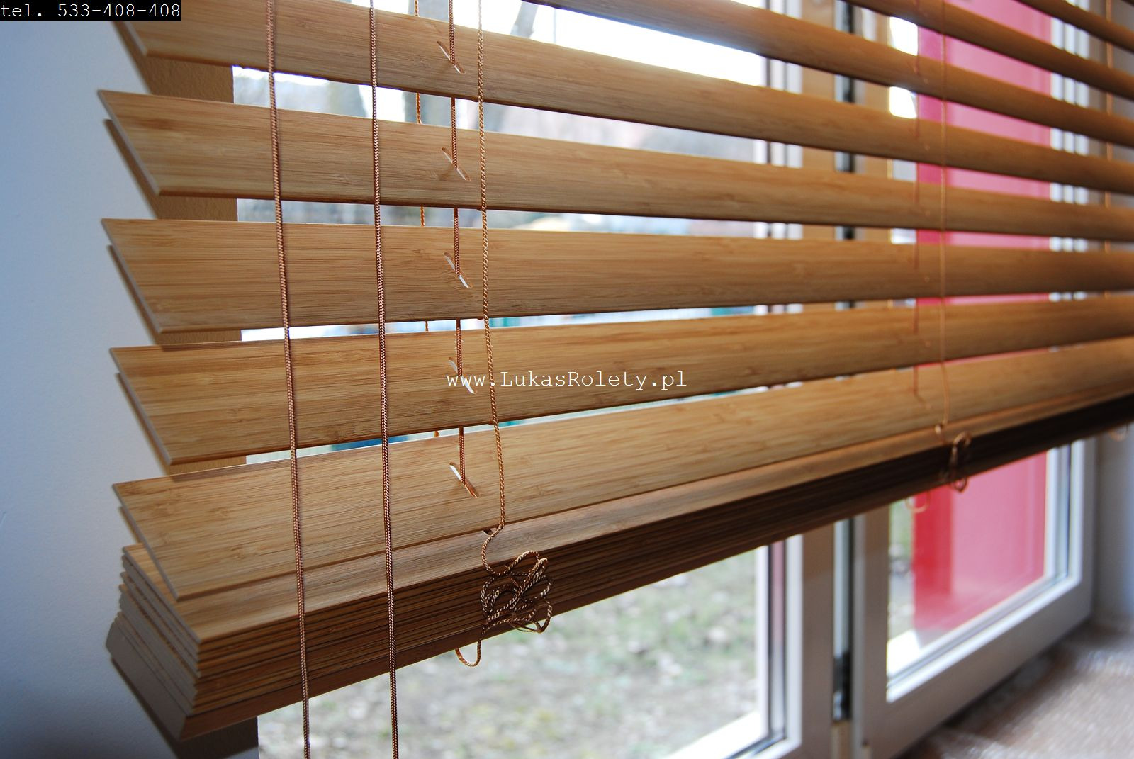 Drewniane lamele w poziomych żaluzjach z widocznymi sznurkami sterującymi, zamontowane w oknie z widokiem na zewnątrz.