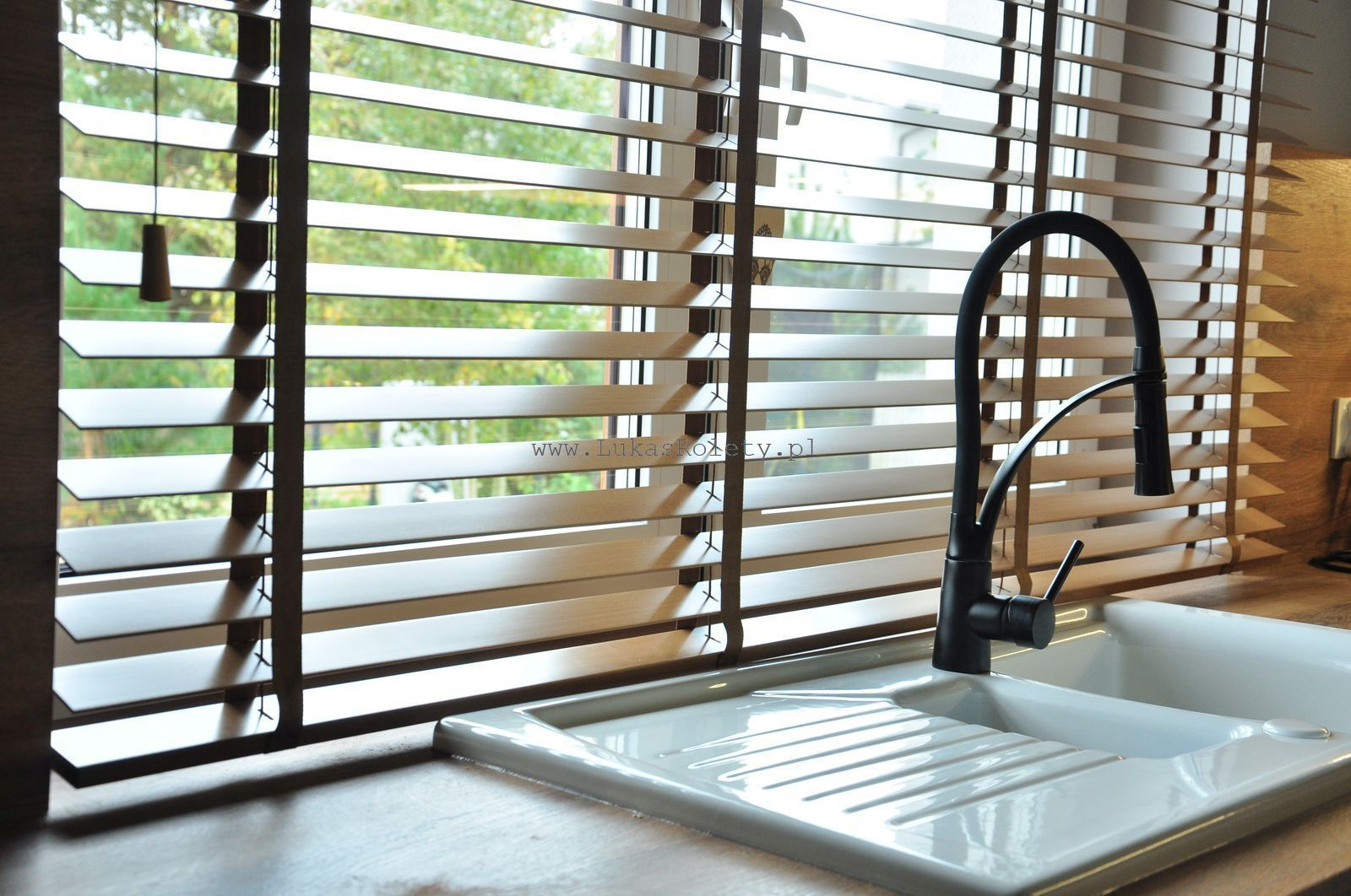 Drewniane żaluzje poziome, widoczne z perspektywy zlewu kuchennego, z zielenią drzew za oknem.