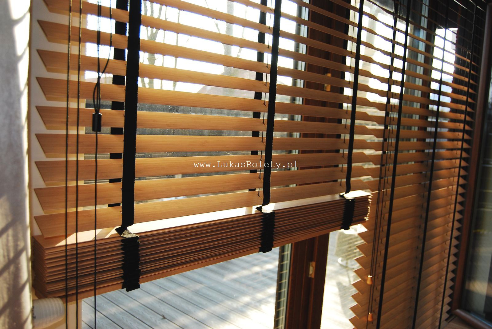 Drewniane lamele w kolorze jasnego drewna, połączone czarnymi taśmami, tworzące osłonę okienną przy dużym oknie z widokiem na zewnątrz.