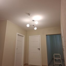 RECO-BUD - Korytarz w trakcie malowania: beżowe ściany, białe drzwi, puszki z farbą na podłodze, drabina aluminiowa, nowoczesna lampa sufitowa.