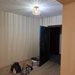 RECO-BUD - Pokój w trakcie prac przygotowawczych: zabezpieczone folią drzwi, wiadro z farbą, butelki z płynami i narzędzia na podłodze z paneli, sufit z lampą w stylu glamour.