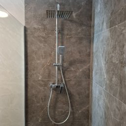 RECO-BUD - Nowoczesny zestaw prysznicowy z deszczownicą i słuchawką na tle ściany wyłożonej płytkami imitującymi marmur w odcieniach szarości i brązu, widoczny fragment szklanej kabiny.