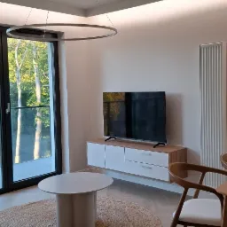 Minimalistyczny salon w Kołobrzegu: okrągły dywan, stolik, szafka RTV i telewizor. Duże okno z widokiem na zieleń. Nowoczesne oświetlenie sufitowe i grzejnik.