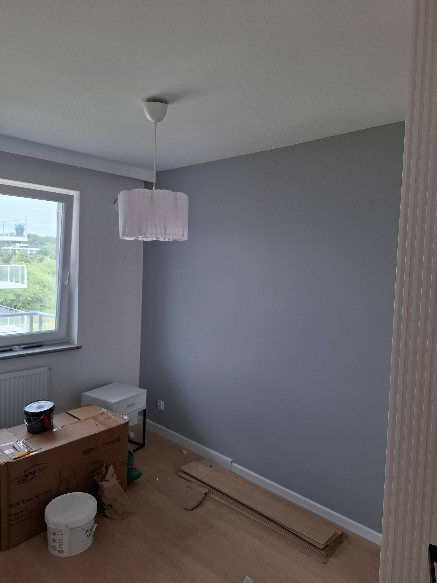 Świeżo pomalowane na szaro pomieszczenie z widocznymi puszkami farby, kartonem po meblach i panelami podłogowymi czekającymi na montaż, oświetlone przez wiszącą lampę z abażurem i światło z okna.