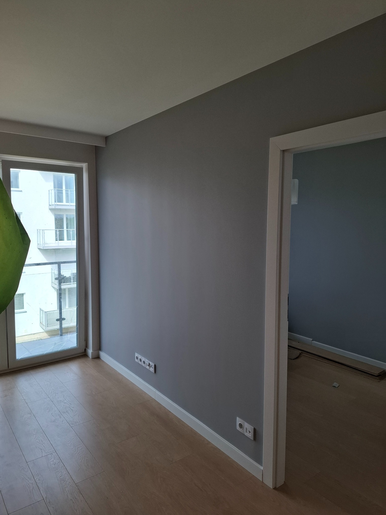 Pomieszczenie po malowaniu na szaro w Kołobrzegu, widoczne gładkie ściany, drewniana podłoga i białe listwy przypodłogowe, z oknem balkonowym i otwartymi drzwiami do kolejnego pokoju.