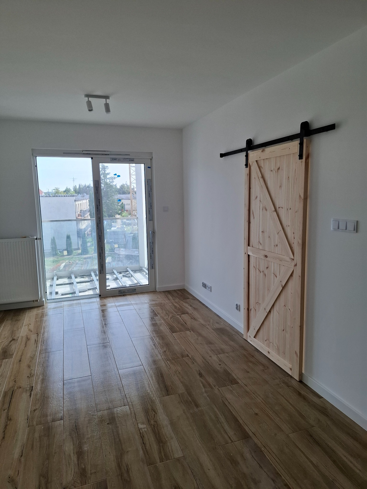 Jasne drewniane drzwi przesuwne w stylu rustykalnym, zamontowane na czarnej szynie, widoczne w nowocześnie wykończonym wnętrzu z panelami podłogowymi imitującymi drewno.