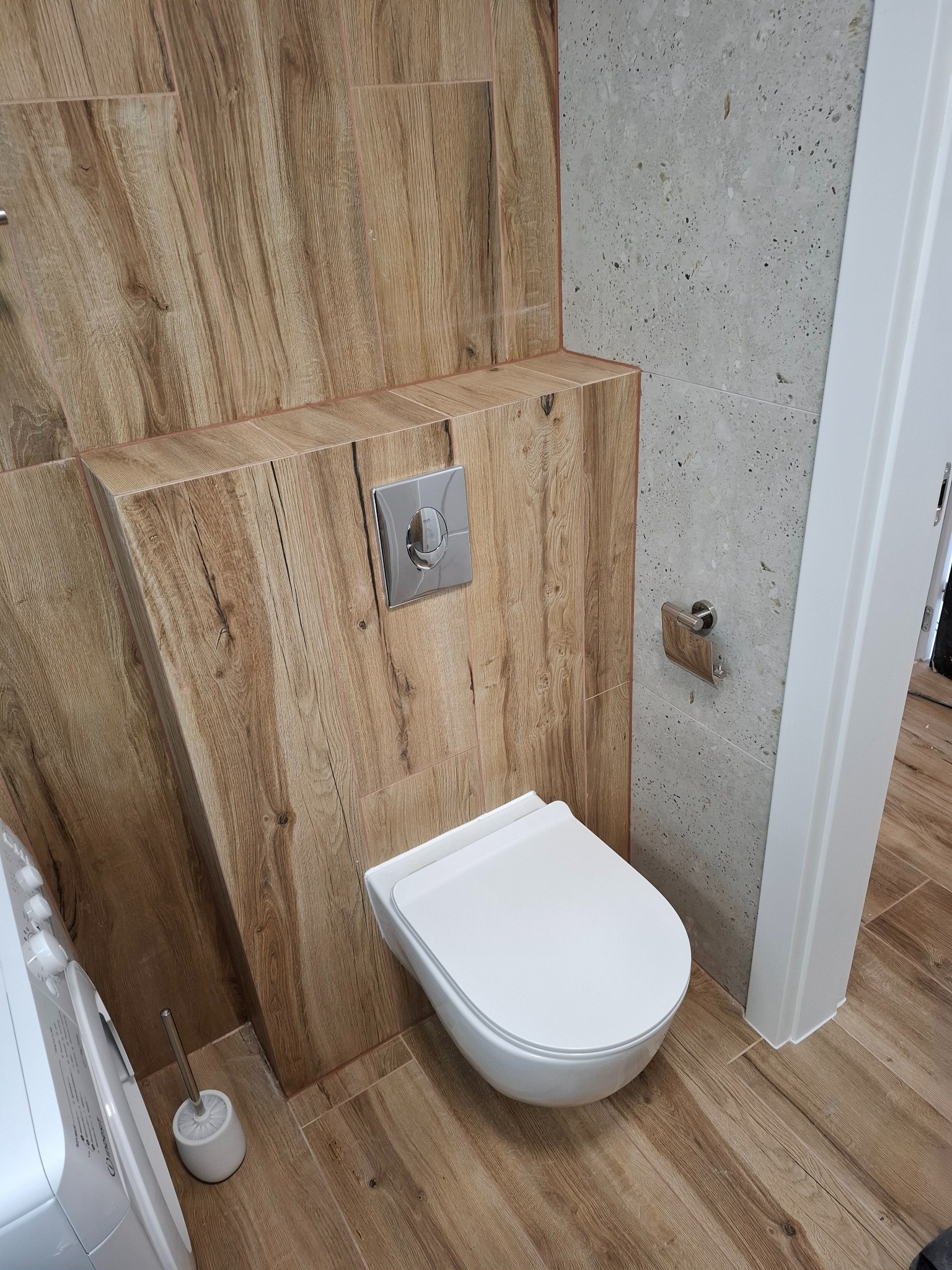 Nowoczesna toaleta w Kołobrzegu z drewnopodobnymi płytkami na ścianie i podłodze, kontrastującymi z szarą ścianą z fakturą i białym sanitariatem.