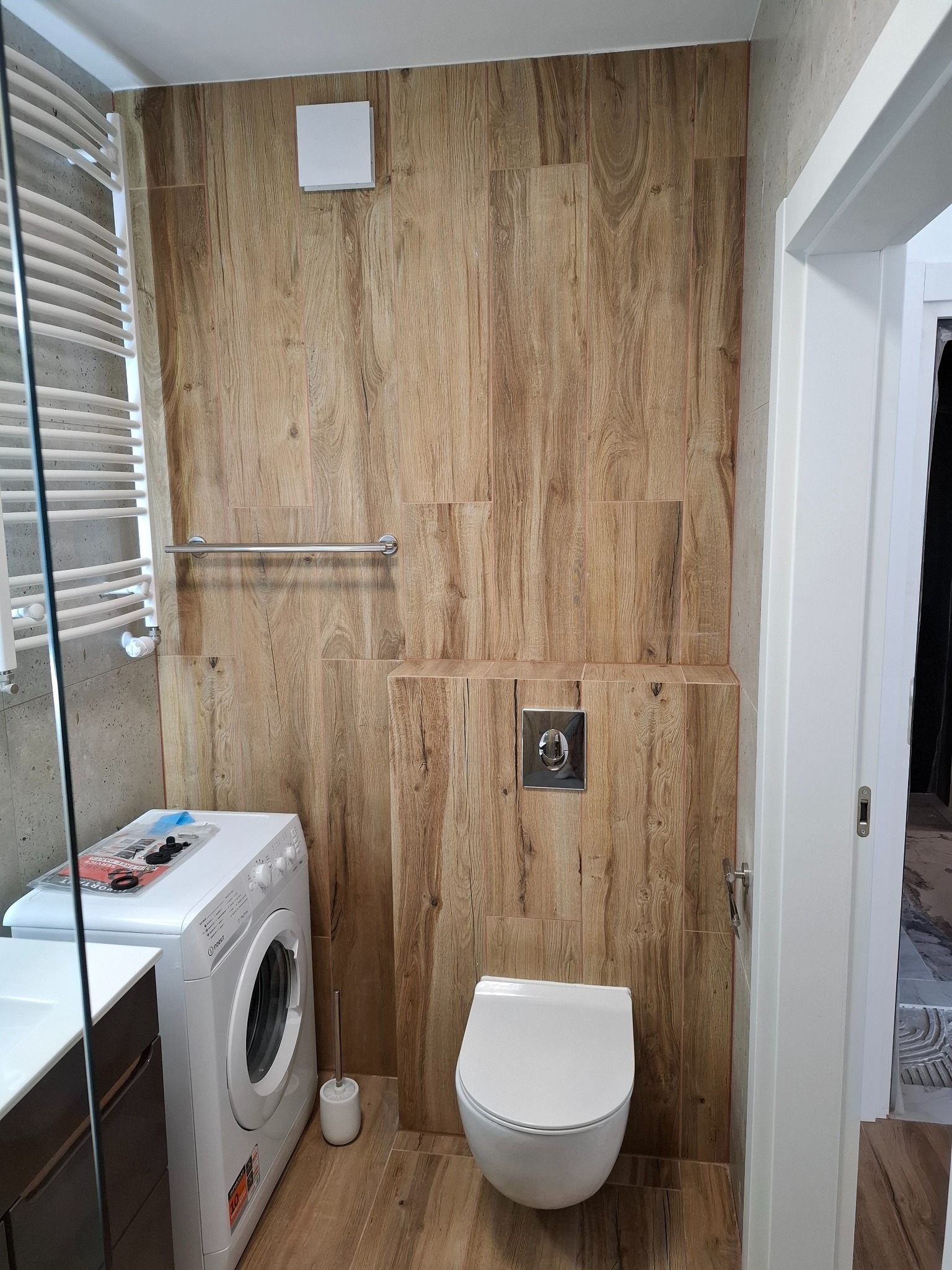 Nowoczesna łazienka z drewnopodobnymi płytkami na ścianach i podłodze, wisząca toaleta, biała pralka i grzejnik drabinkowy.