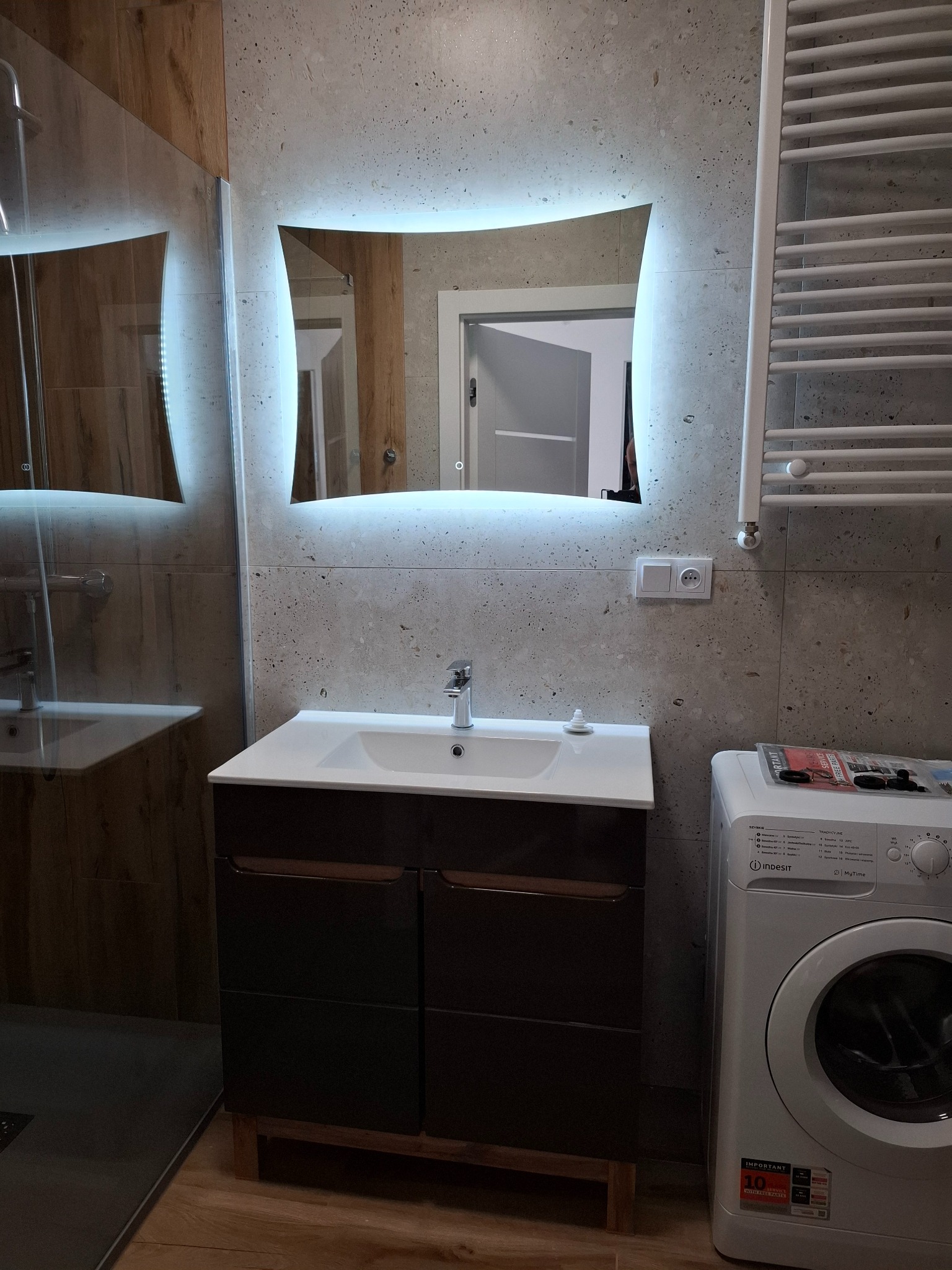Nowoczesna łazienka z podświetlanym lustrem, umywalką z szafką w ciemnym drewnie, grzejnikiem drabinkowym i pralką Indesit, wykończona płytkami imitującymi kamień.