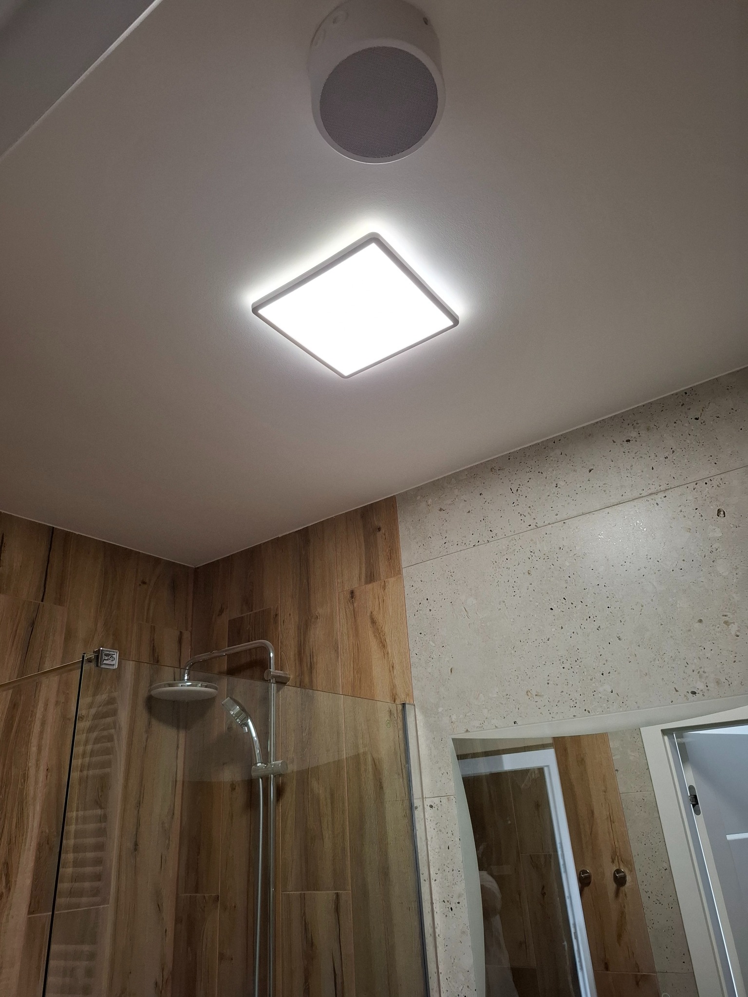 Nowoczesna łazienka z drewnopodobnymi płytkami, kabiną prysznicową ze szklaną ścianką, kwadratową lampą sufitową LED i okrągłym głośnikiem w suficie.