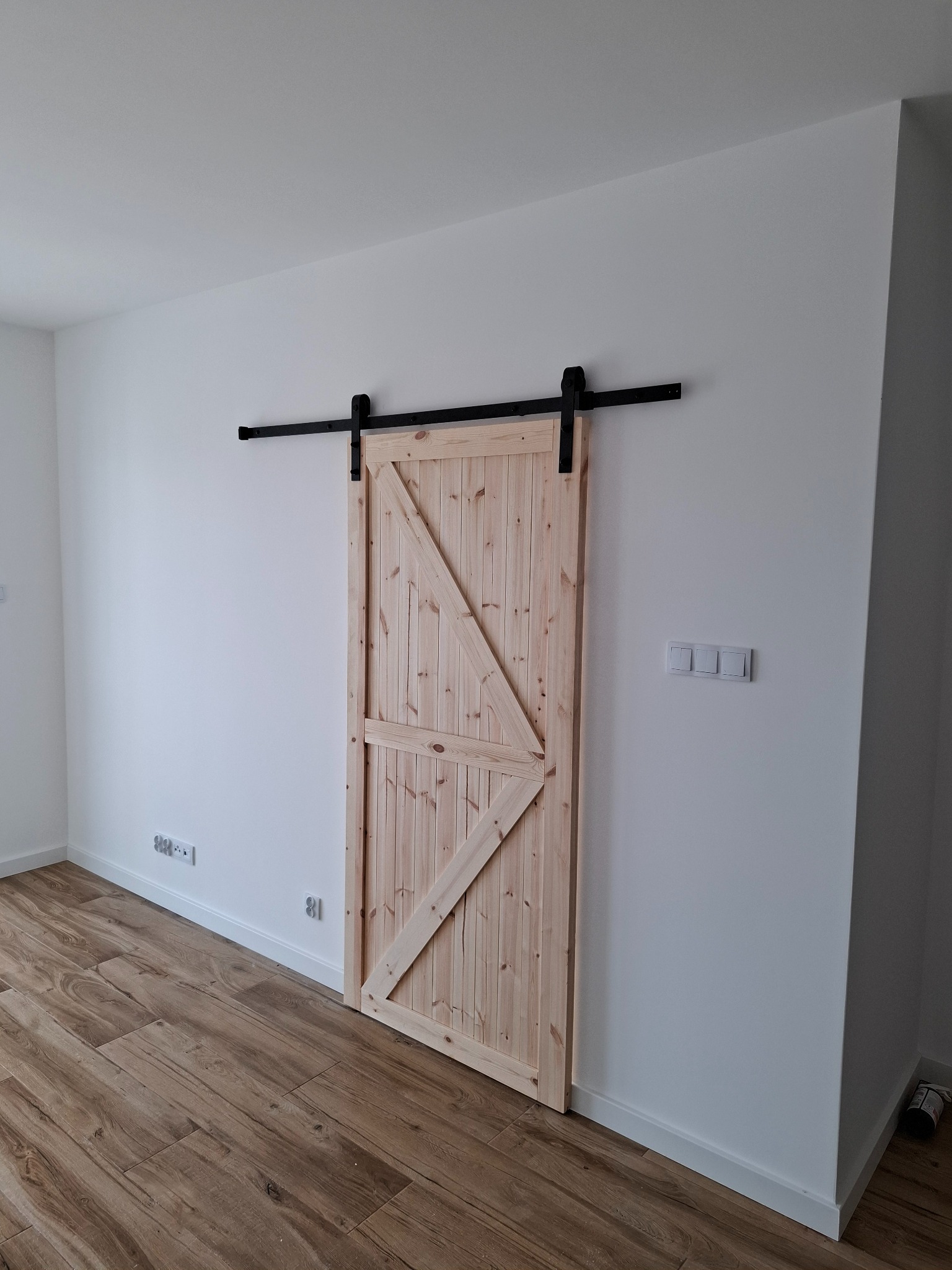 Drewniane drzwi przesuwne w stylu rustykalnym zamontowane na czarnej szynie, na tle białej ściany z widocznymi gniazdkami elektrycznymi i włącznikami światła, podłoga wyłożona panelami imitującymi...