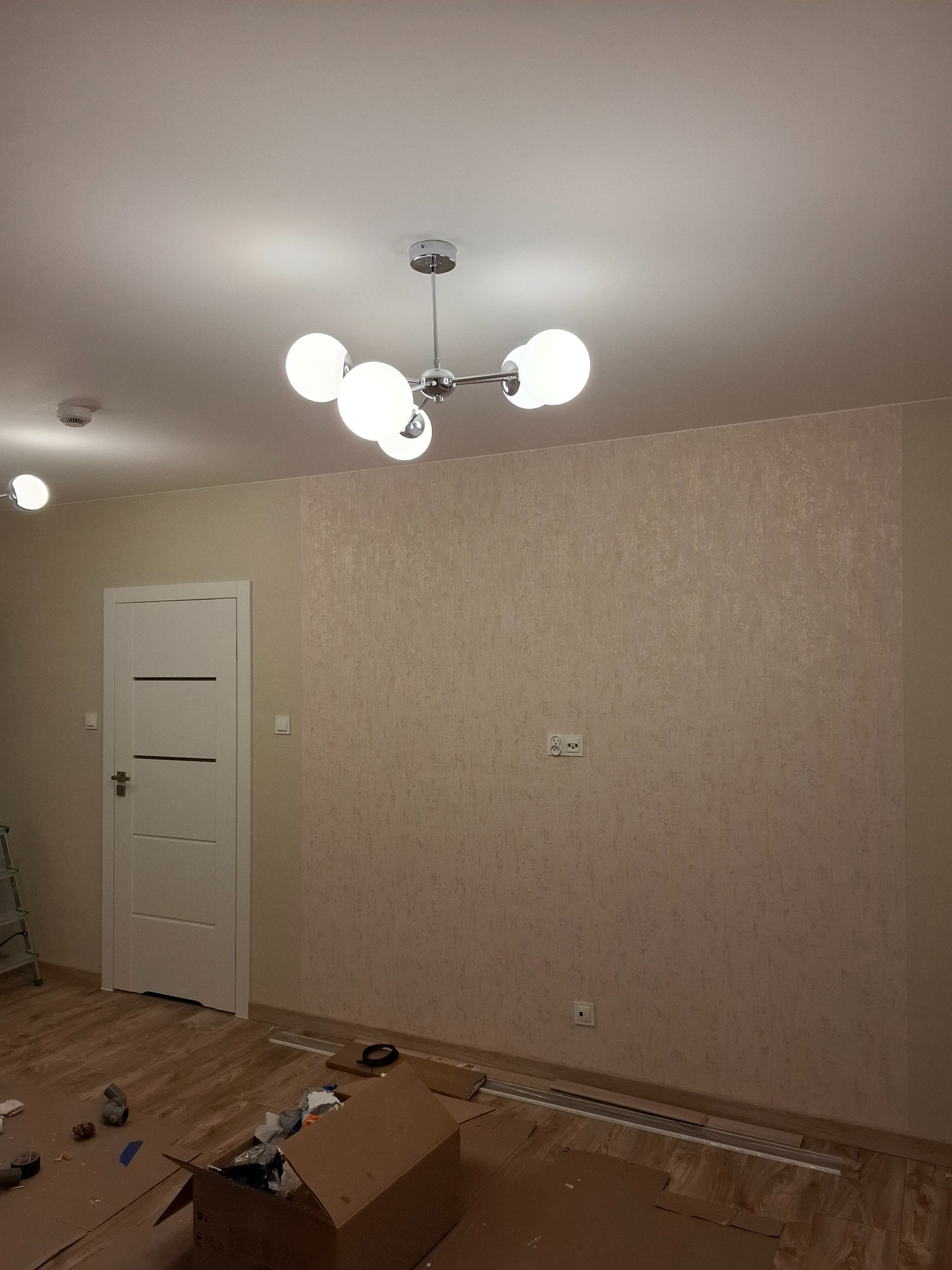 Pokój po remoncie z nową tapetą o delikatnym wzorze, nowoczesną lampą sufitową z kulistymi kloszami i widoczną białą stolarką drzwiową. Na podłodze widoczne ślady po pracach wykończeniowych...