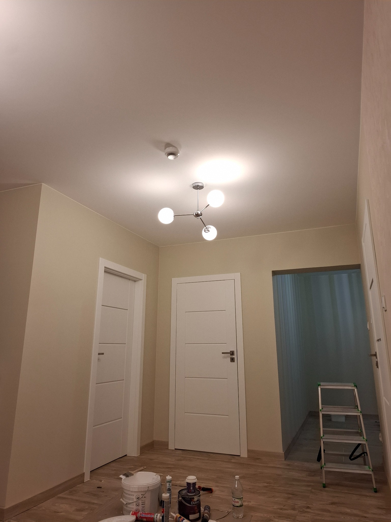 Korytarz w trakcie malowania: beżowe ściany, białe drzwi, puszki z farbą na podłodze, drabina aluminiowa, nowoczesna lampa sufitowa.