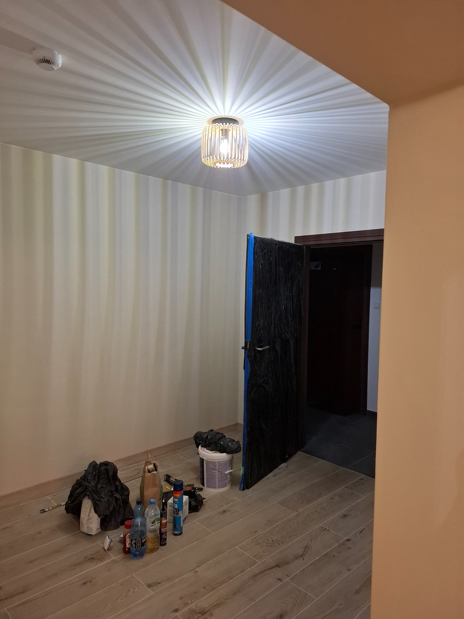 Pokój w trakcie prac przygotowawczych: zabezpieczone folią drzwi, wiadro z farbą, butelki z płynami i narzędzia na podłodze z paneli, sufit z lampą w stylu glamour.