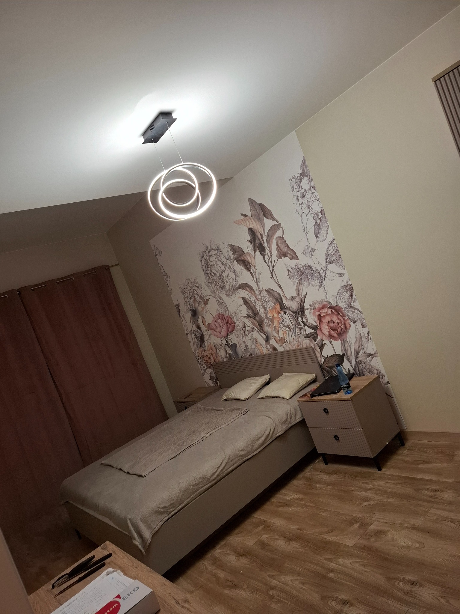 Sypialnia z łóżkiem jednoosobowym, szafką nocną, lampą wiszącą z okręgów LED i tapetą w kwiaty na ścianie akcentującej.