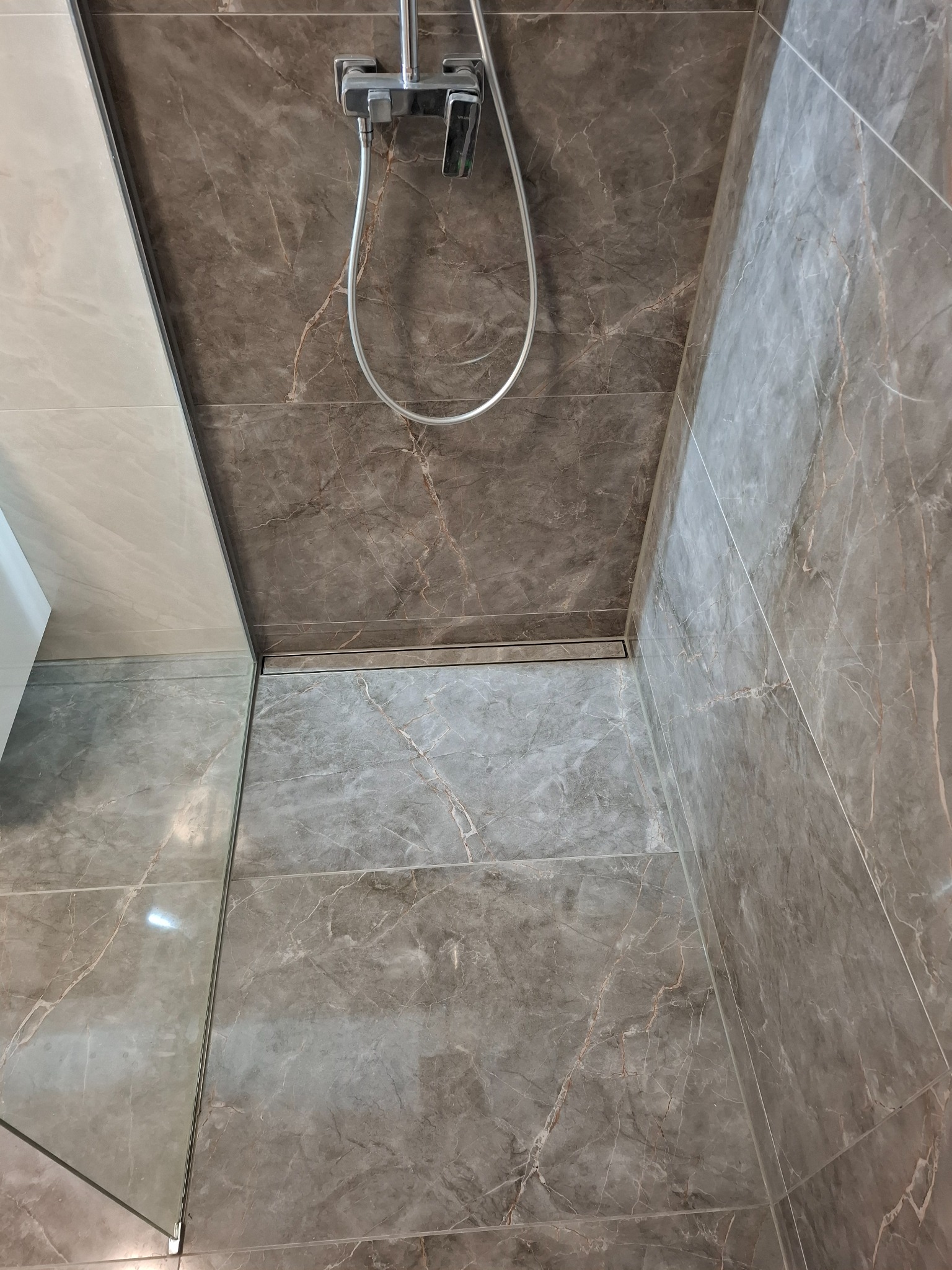 Wykończona kabina prysznicowa z szarymi płytkami imitującymi marmur, widoczny odpływ liniowy i bateria prysznicowa ze słuchawką.