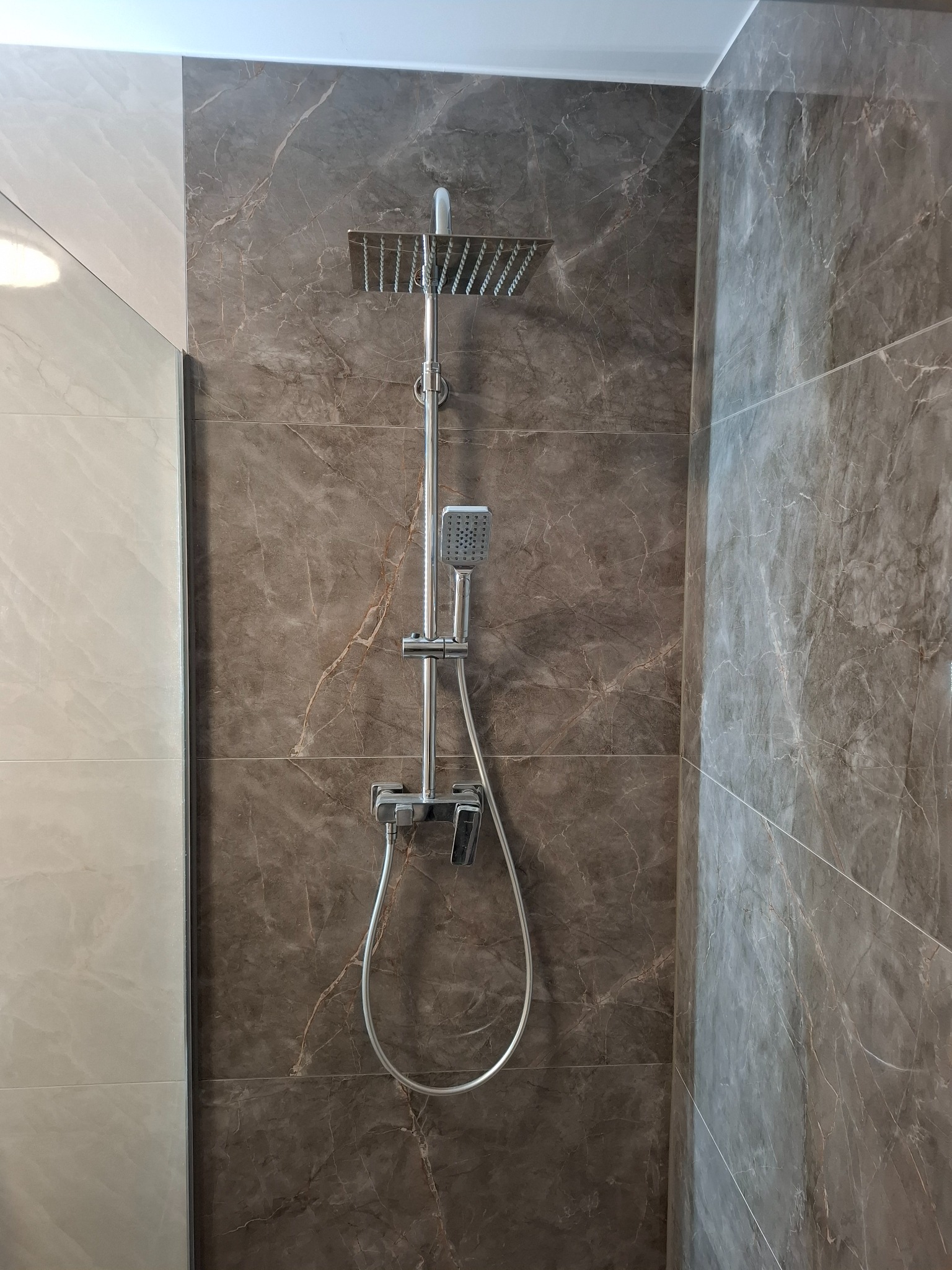 Nowoczesny zestaw prysznicowy z deszczownicą i słuchawką na tle ściany wyłożonej płytkami imitującymi marmur w odcieniach szarości i brązu, widoczny fragment szklanej kabiny.