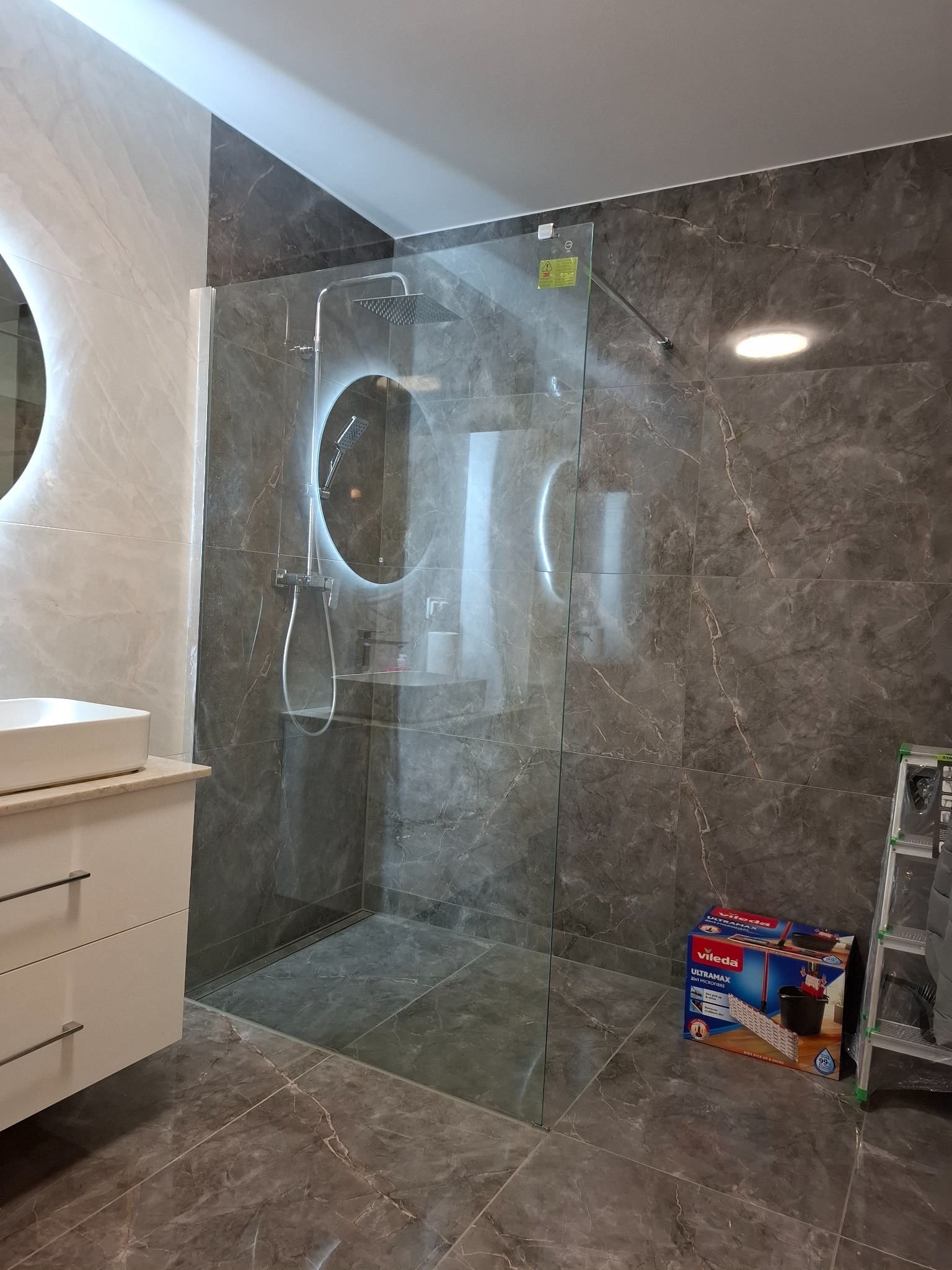 Nowoczesna łazienka z szarymi marmurkowymi płytkami, przeszkloną kabiną prysznicową z okrągłym podświetlanym lustrem, białą umywalką z szafką i drabiną oraz pudełkiem mopa Vileda w rogu.