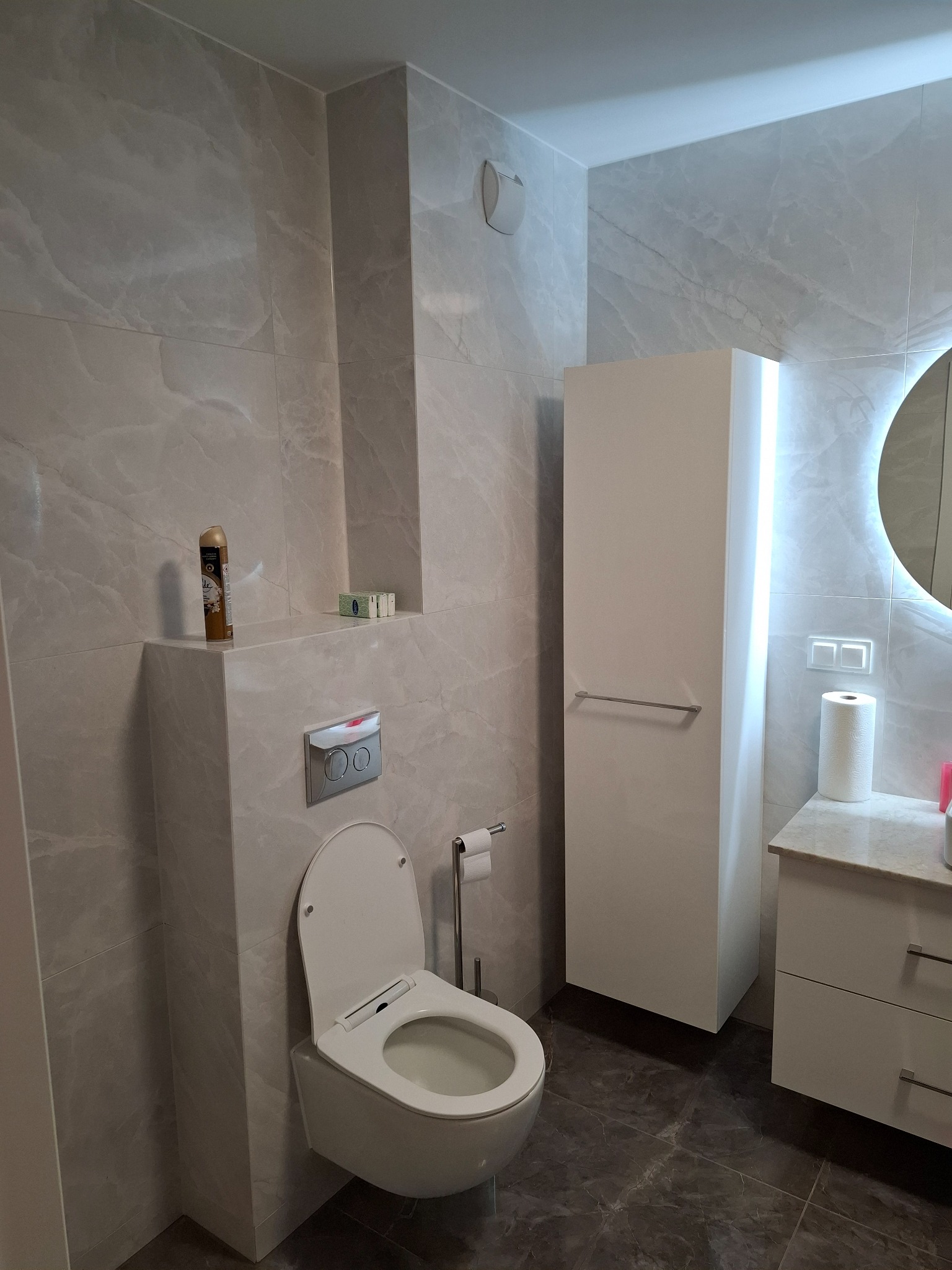 Wykończona łazienka w odcieniach szarości z marmurkową glazurą, podwieszana toaleta z zabudową, biała szafka z oświetleniem LED i marmurowym blatem, oraz wysoka szafka z relingiem na ręcznik.