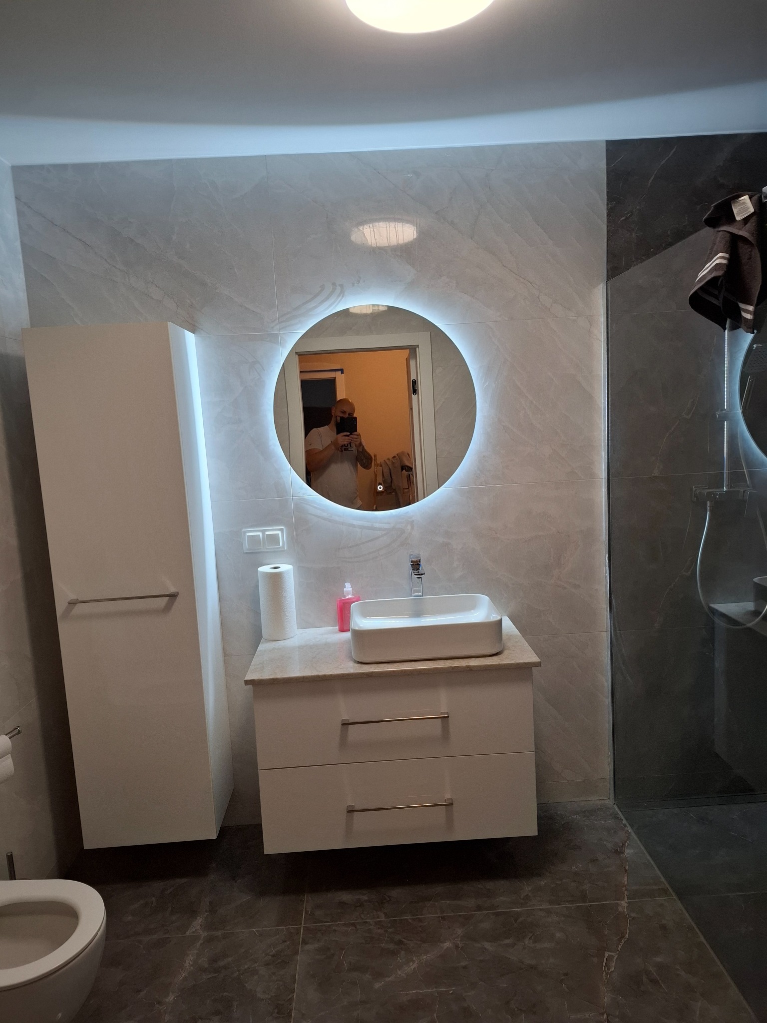 Nowoczesna łazienka z umywalką nablatową, podświetlanym okrągłym lustrem i szarymi płytkami imitującymi marmur, widoczna kabina prysznicowa ze szklaną ścianką.