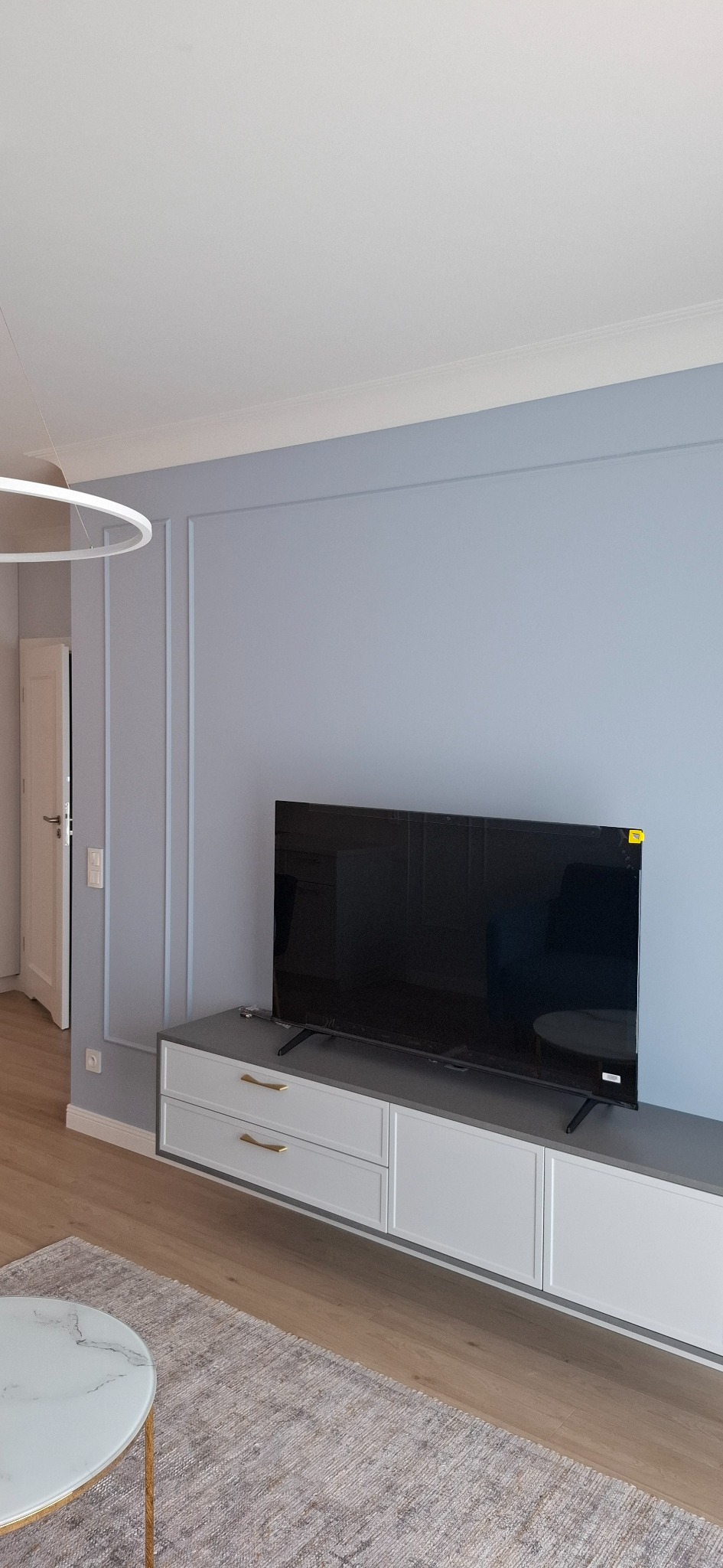 Pokój z telewizorem na szafce RTV, ściana w delikatnym błękicie z dekoracyjnymi listwami. Widoczny fragment dywanu i stolika kawowego.