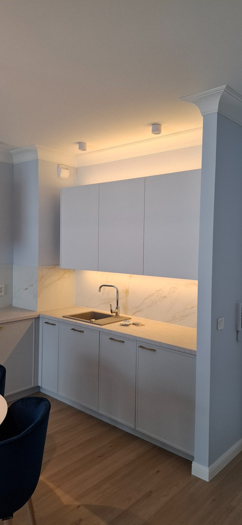 Nowoczesna kuchnia z białymi szafkami, marmurowym blatem i subtelnym oświetleniem LED pod szafkami. Widoczny fragment jadalni z krzesłem.