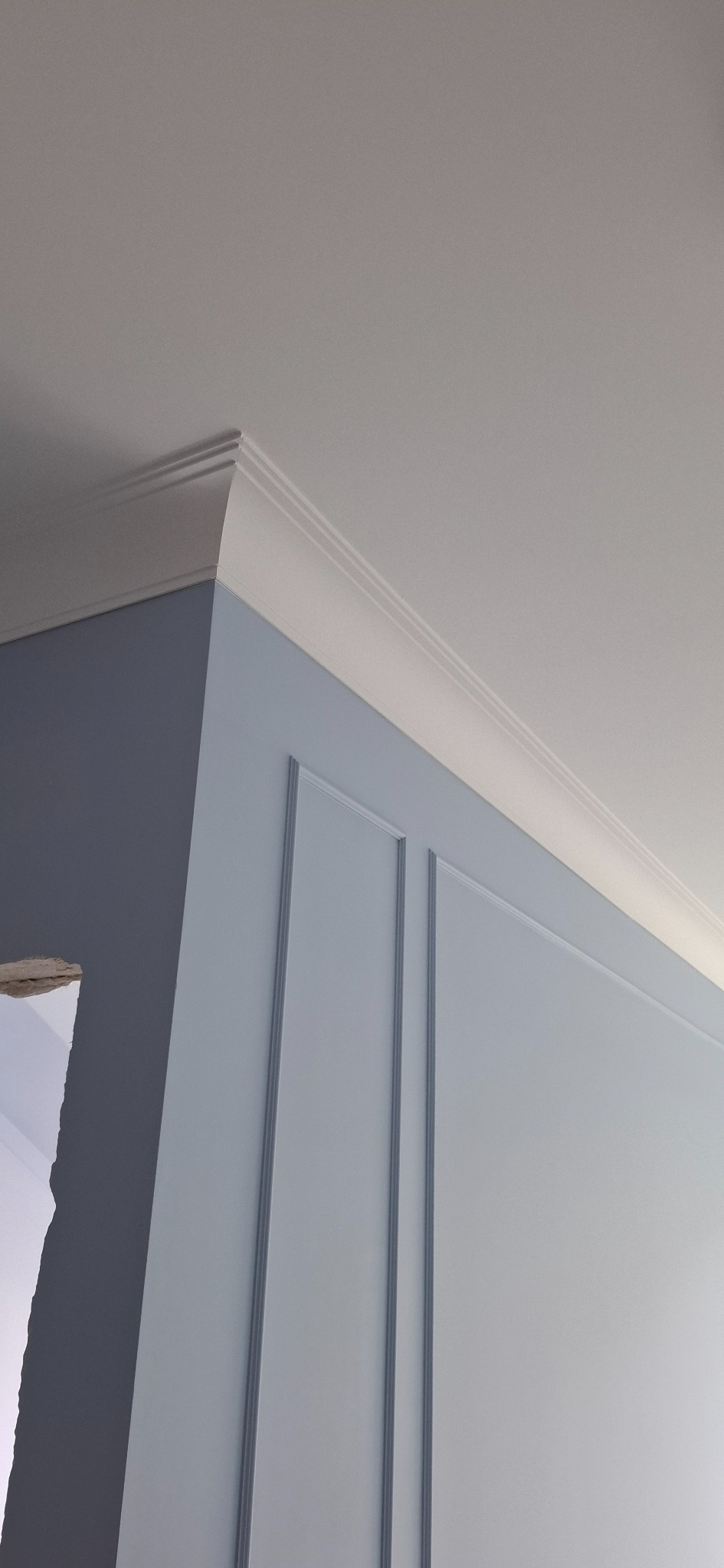 Jasnoszara ściana z dekoracyjnymi listwami i białym sztukateryjnym wykończeniem sufitu, widok z dołu. Fragment ściany w ciemniejszym odcieniu szarości po lewej.