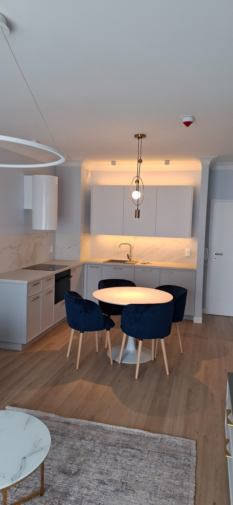 Jasna kuchnia z marmurowym blatem i okrągłym stołem otoczonym granatowymi krzesłami. Nad stołem designerska lampa. Nowoczesne wykończenie wnętrza.