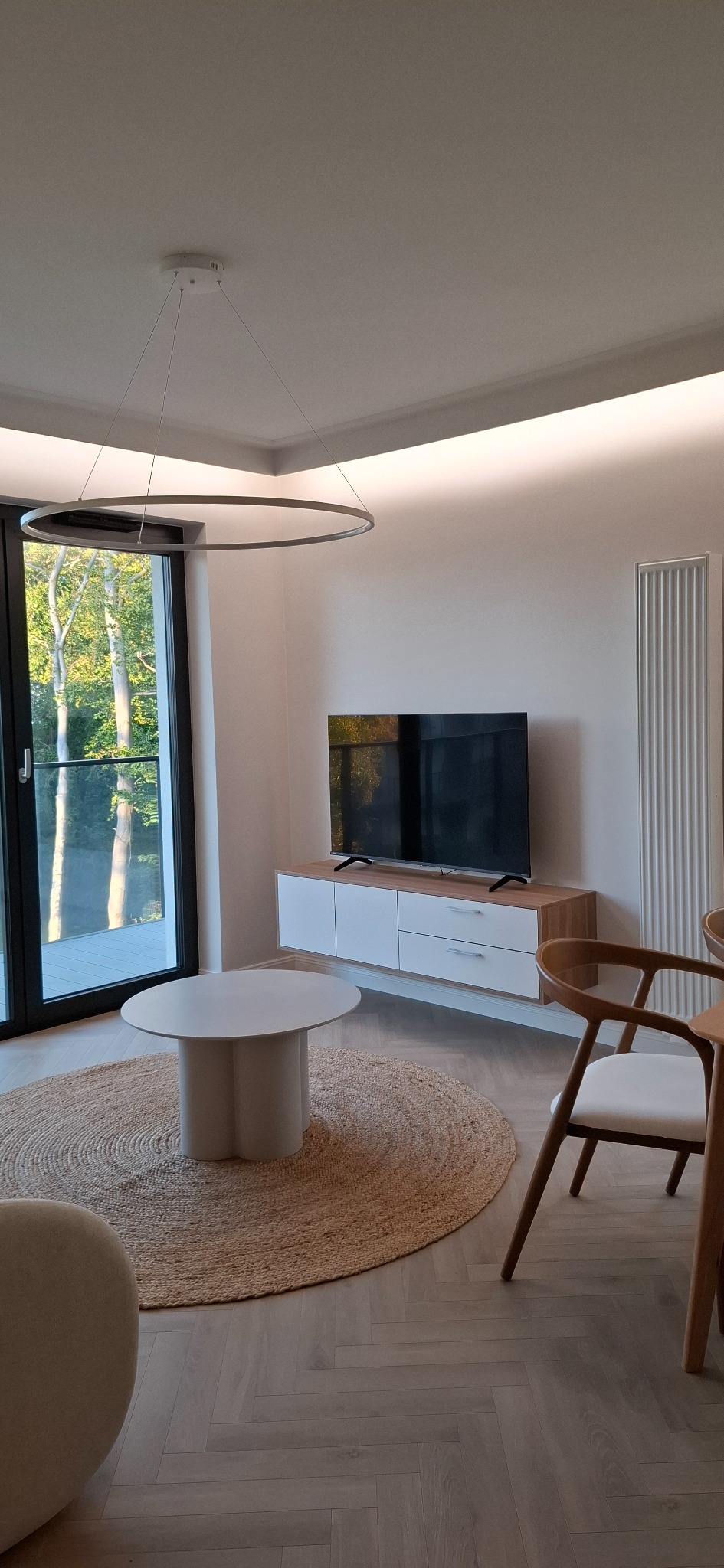 Minimalistyczny salon w Kołobrzegu: okrągły dywan, stolik, szafka RTV i telewizor. Duże okno z widokiem na zieleń. Nowoczesne oświetlenie sufitowe i grzejnik.