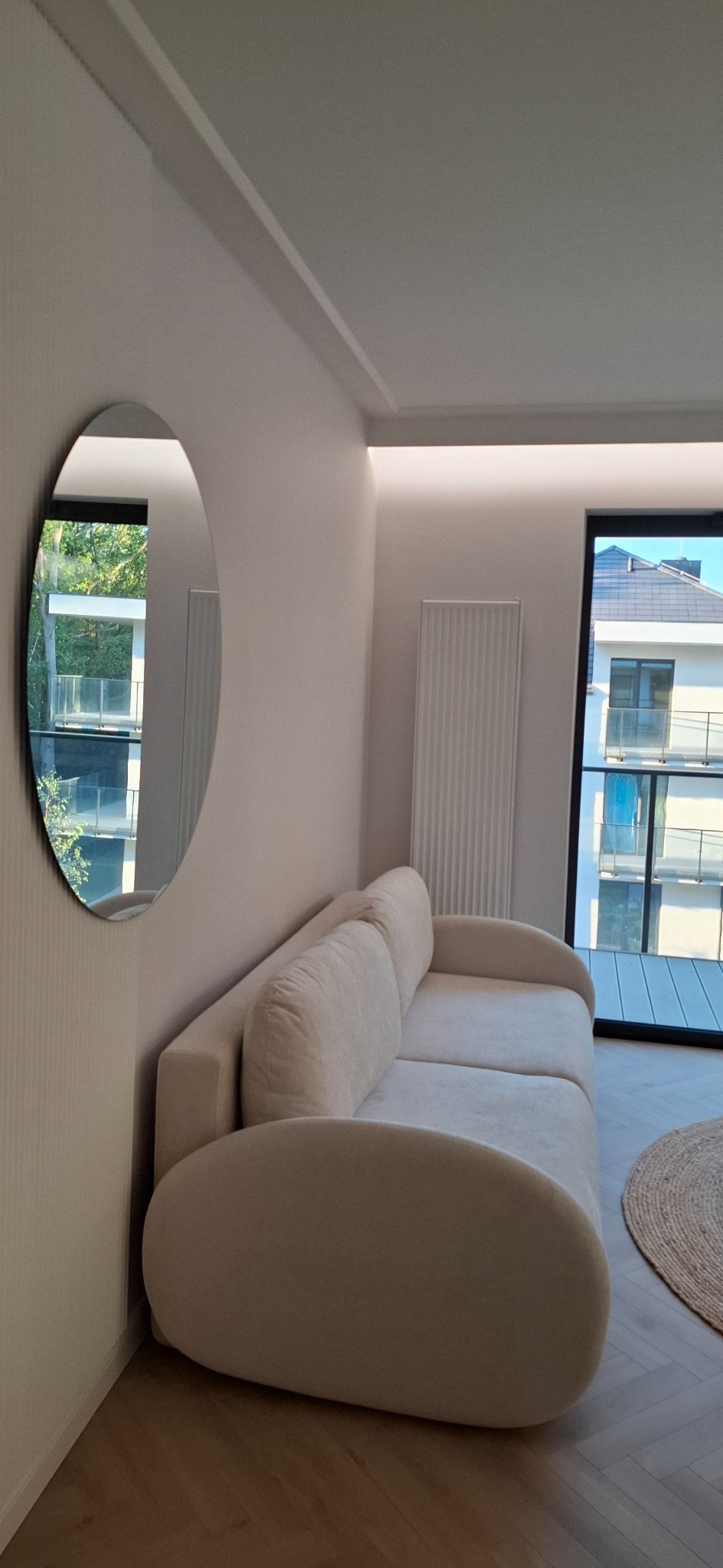 Jasny salon z beżową sofą, okrągłym lustrem i grzejnikiem panelowym na ścianie. Widok na budynek z balkonami przez okno. Minimalistyczny wystrój.