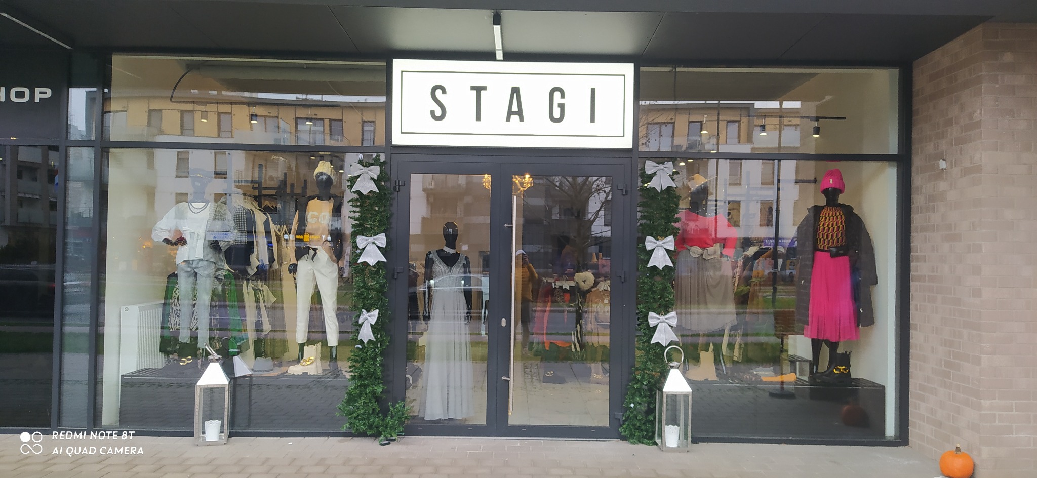 Elegancka witryna butiku odzieżowego 'STAGI' z manekinami prezentującymi różnorodne stylizacje, udekorowana girlandą z białymi kokardami, lampionami i dynią.