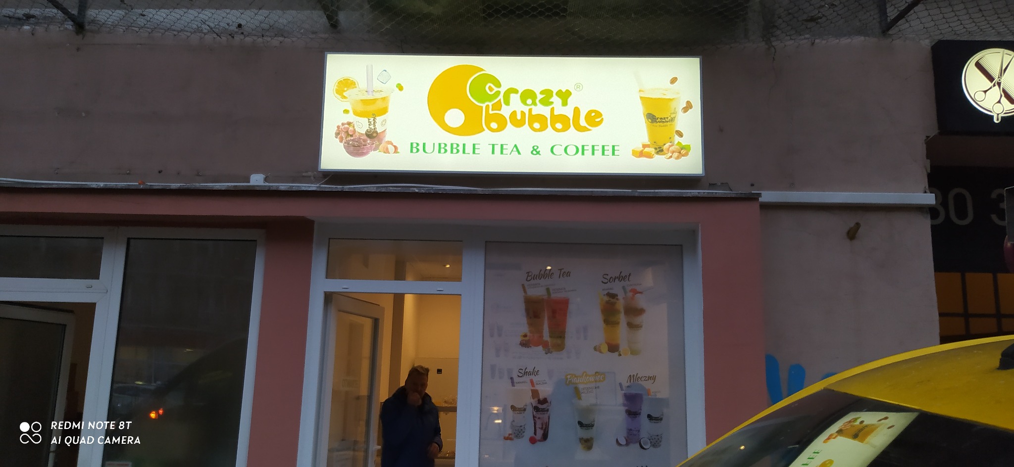 Oświetlony szyld kawiarni 'Crazy Bubble' z grafikami napojów Bubble Tea i Coffee, umieszczony nad wejściem do lokalu. W oknie widoczny mężczyzna.
