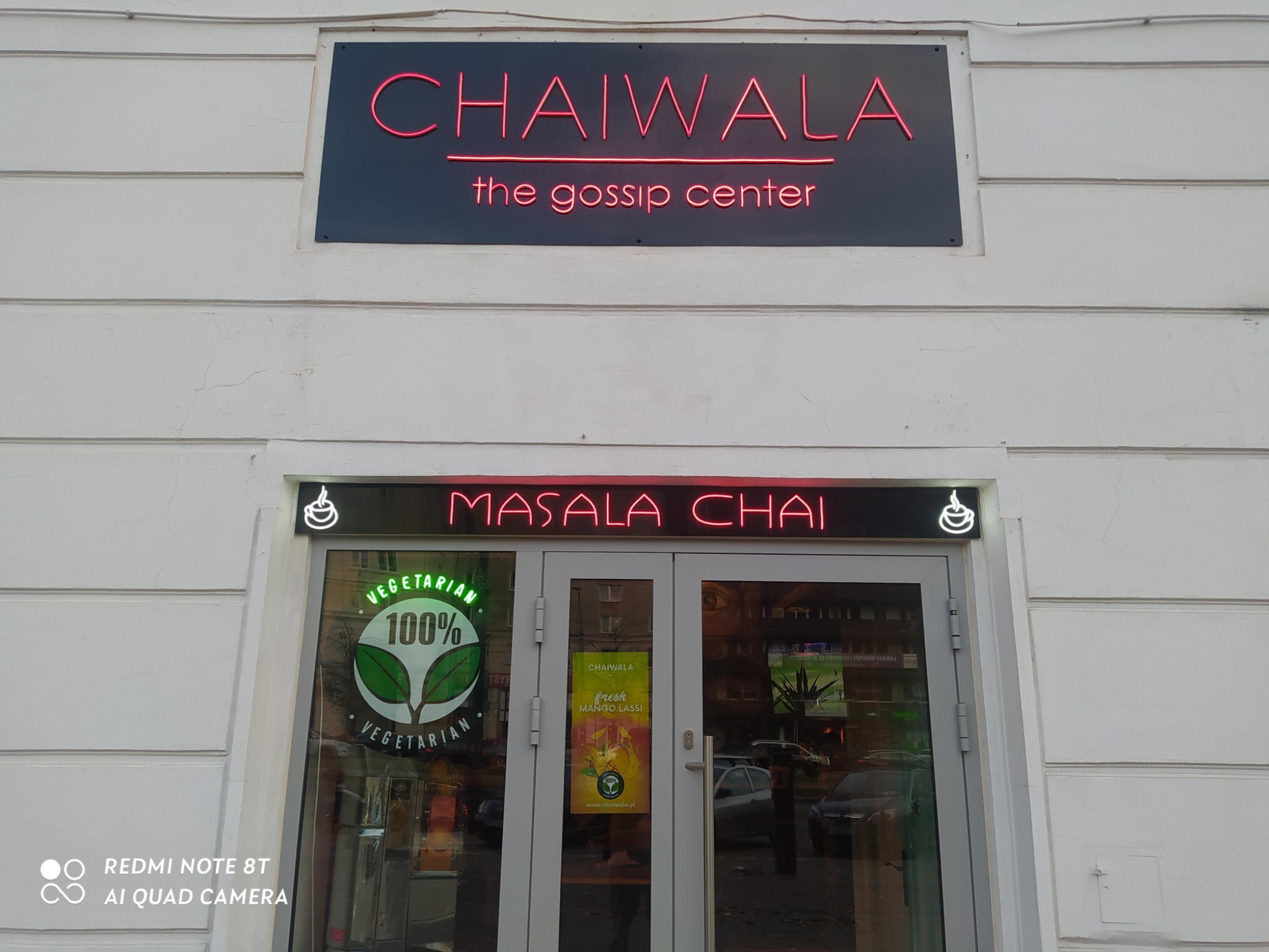 Fasada lokalu Chaiwala z neonowymi szyldami 'Chaiwala the gossip center' i 'Masala Chai' nad wejściem, z naklejką '100% Vegetarian' na szybie.