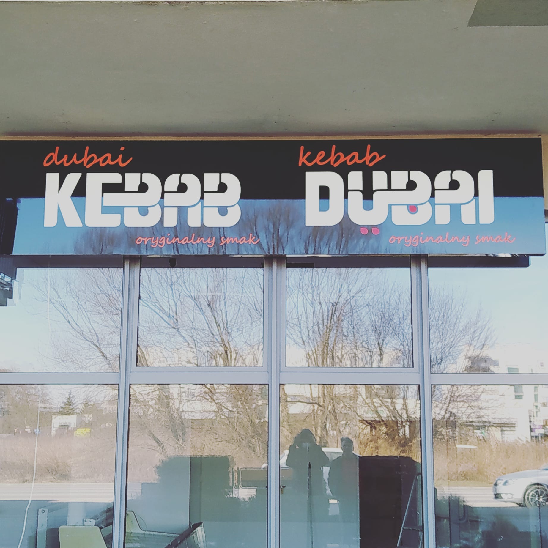 Szyld reklamowy 'Dubai Kebab' z napisem 'oryginalny smak' nad witryną lokalu gastronomicznego, z odbiciem ulicy i przechodniów w szybach.