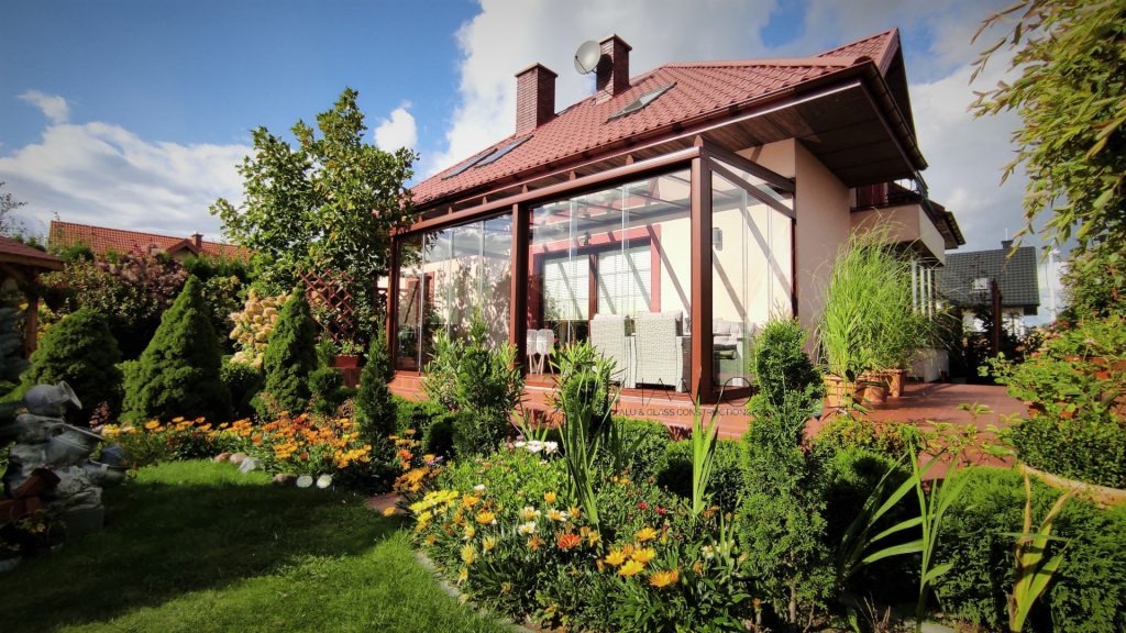 Szklana zabudowa tarasu przylegająca do domu z czerwonym dachem, otoczona zadbanym ogrodem z kwiatami i krzewami.