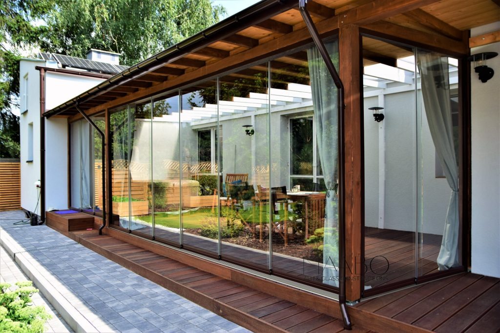 Zadaszony taras z przesuwnymi szklanymi panelami, drewnianą podłogą i widokiem na ogród z pergolą.