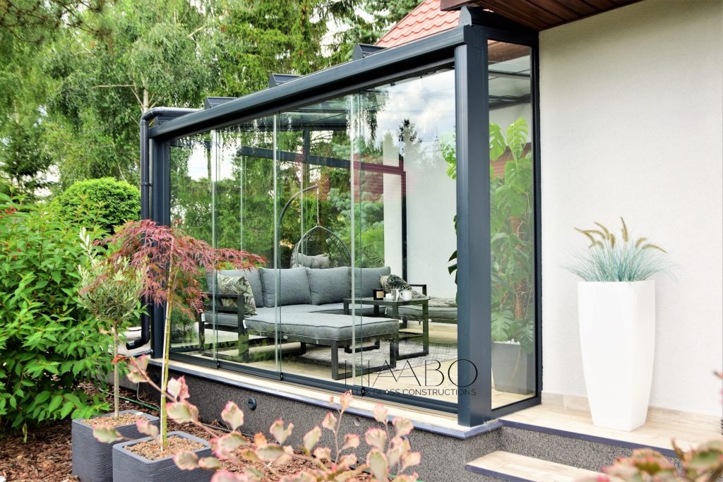 Zadaszony taras z szklanymi ścianami i czarną ramą, wyposażony w szarą sofę i stolik kawowy, widok z ogrodu z ozdobnymi drzewkami w donicach.