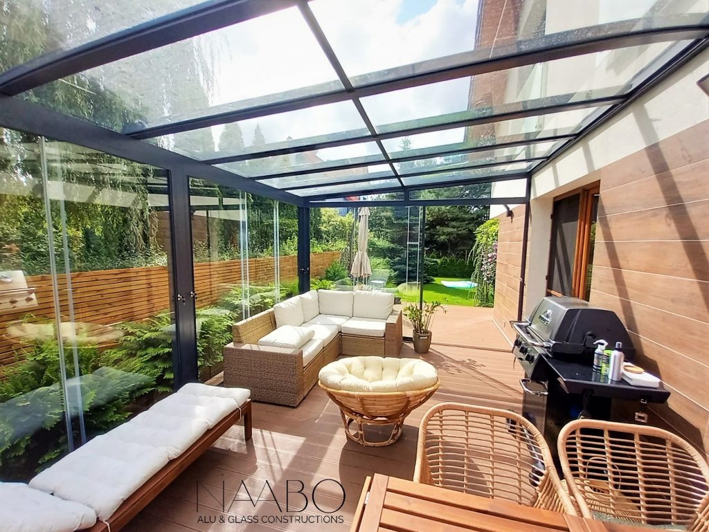 Zadaszony taras z szklanymi ścianami, wyposażony w meble ogrodowe z rattanu, grill i leżankę, widok na ogród z basenem.