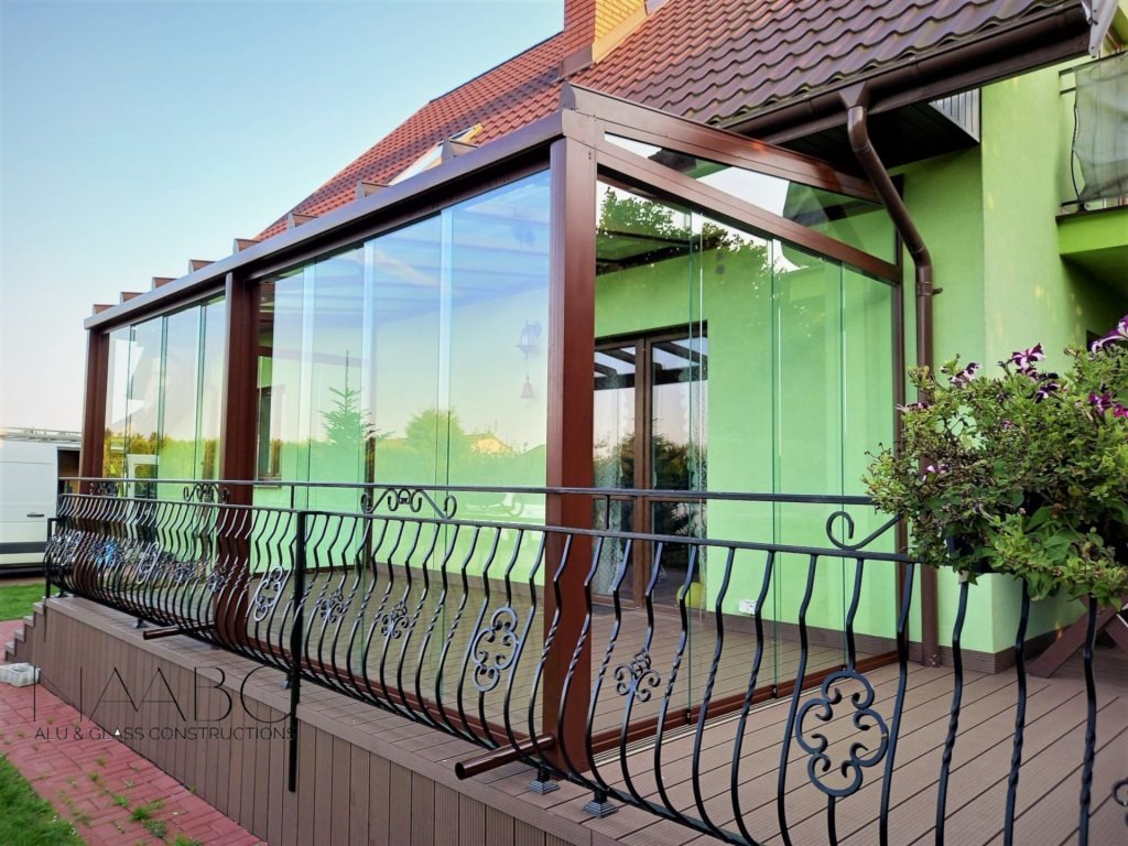 Zadaszony taras z przesuwnymi szklanymi ścianami, brązowa konstrukcja, czarne metalowe balustrady z ozdobnymi elementami, deski tarasowe kompozytowe, zielona ściana domu, częściowo widoczny...