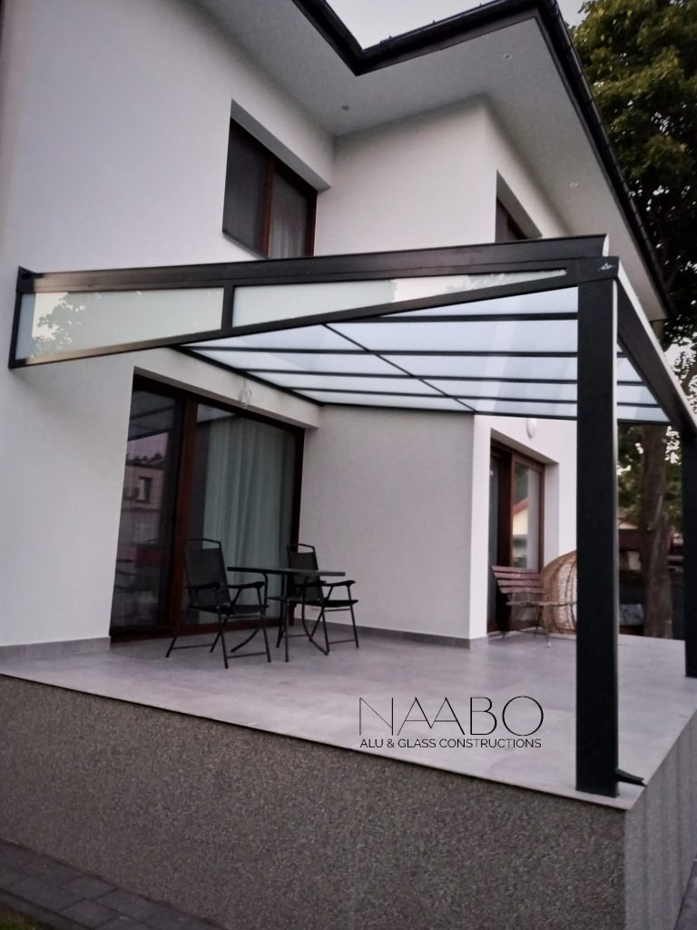 Nowoczesna pergola metalowa z przezroczystym zadaszeniem na tarasie, z minimalistycznym zestawem mebli ogrodowych, widoczny fragment elewacji domu i logo firmy na pierwszym planie.