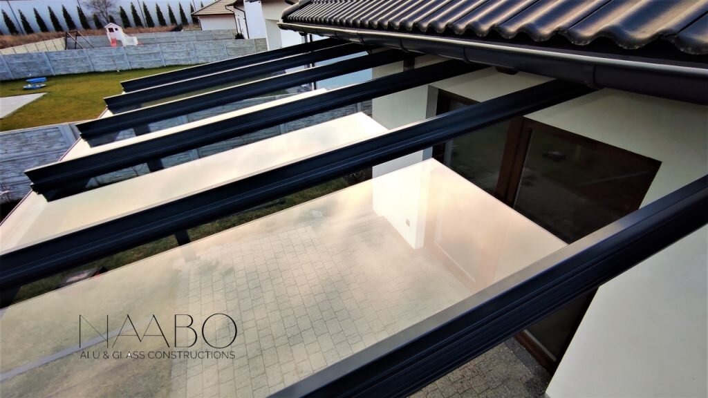 Szklane zadaszenie tarasu z czarną ramą, widok od dołu, na szkle odbija się niebo i otoczenie domu.